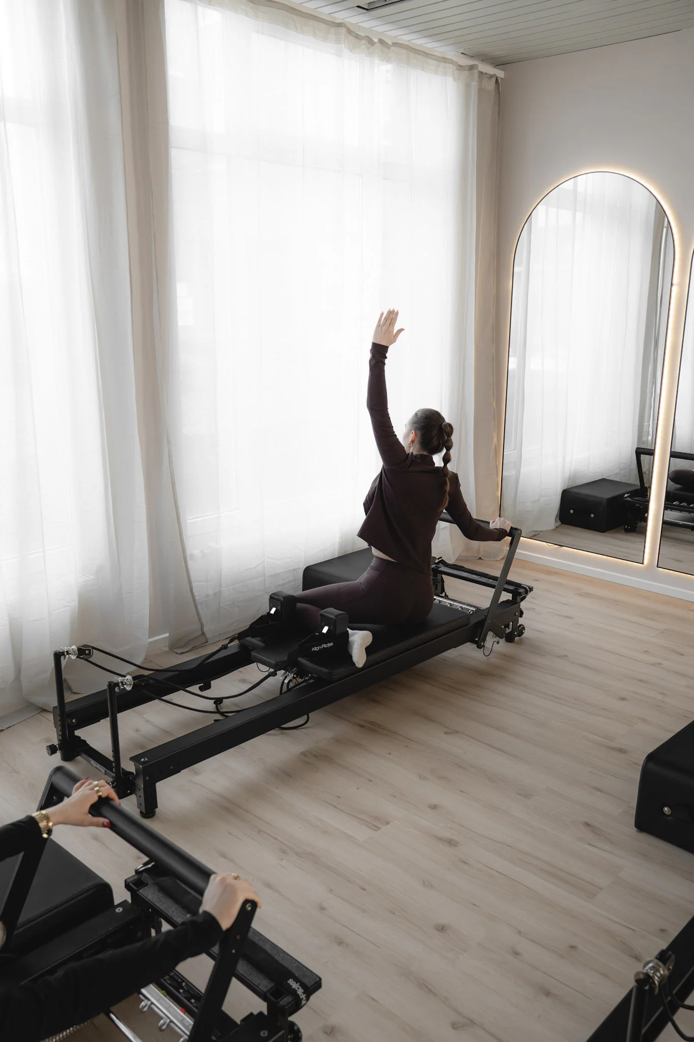 Frau in Sportbekleidung beim Pilates-Training auf einem Reformer-Gerät in einem hellen Raum mit Holzfußboden und großem beleuchtetem Spiegel.