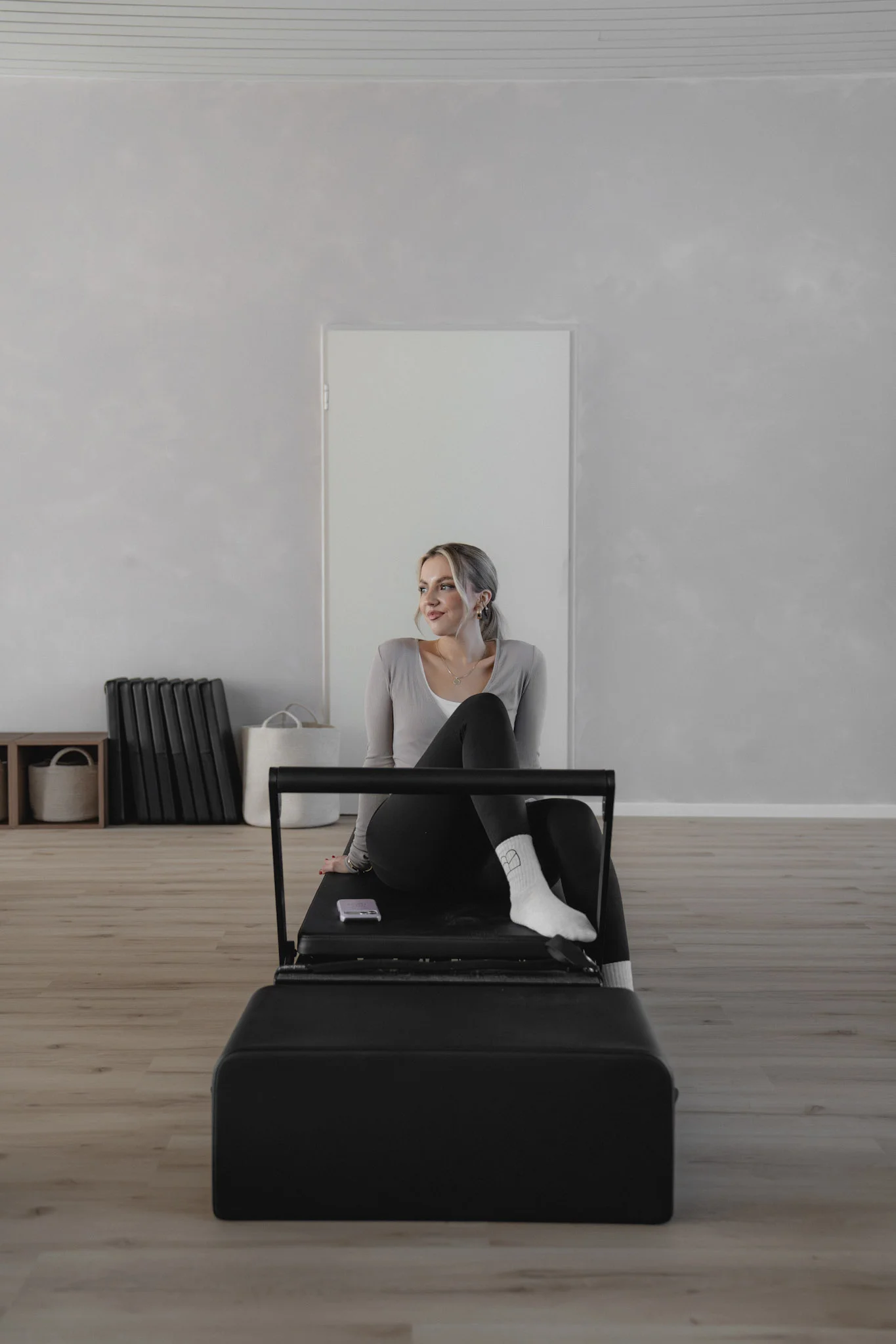 Frau sitzt entspannt auf einem Pilates-Reformergerät in einem hellen, minimalistischen Studio.