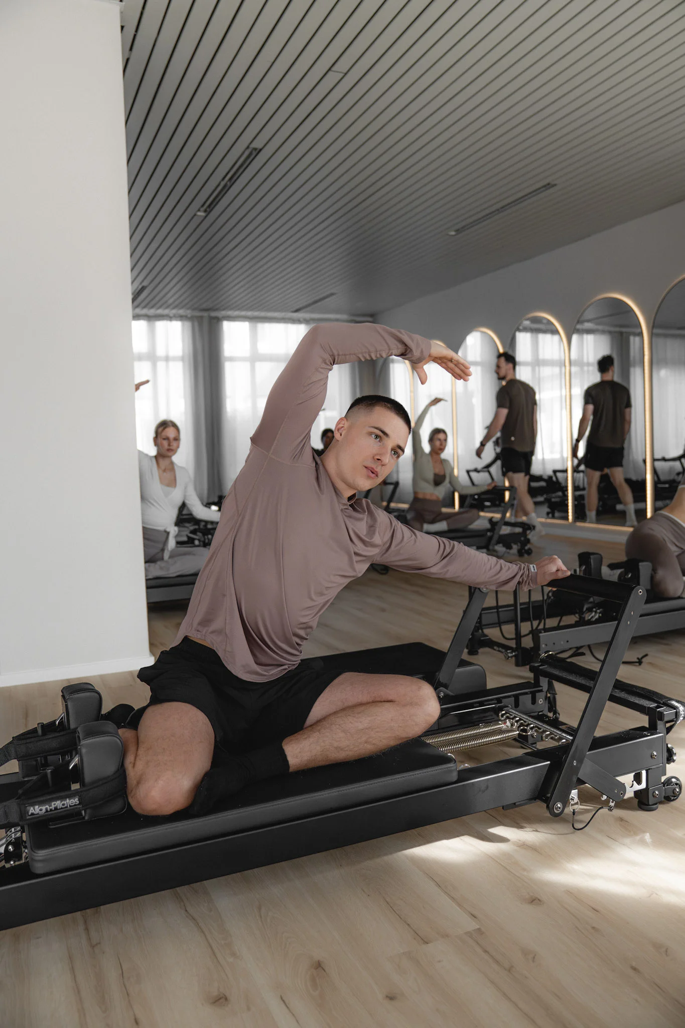 Mann macht seitliche Dehnübung auf einem Pilates-Reformer in einem Plilates-Studio mit Spiegeln im Hintergrund.