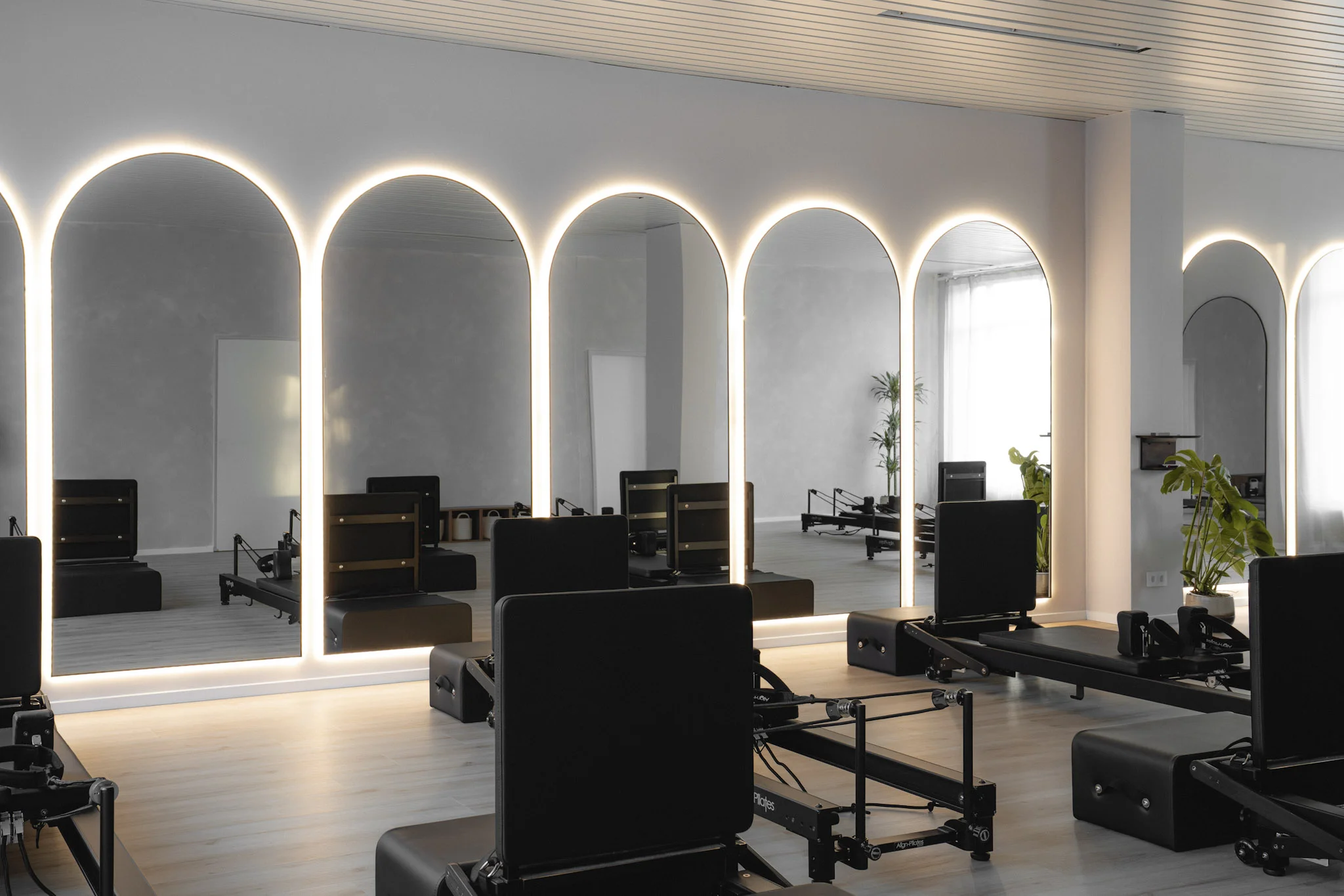 Moderner Pilates-Trainingsraum mit beleuchteten Bogenform-Spiegeln und schwarzen Pilates-Reformern auf hellem Holzboden.