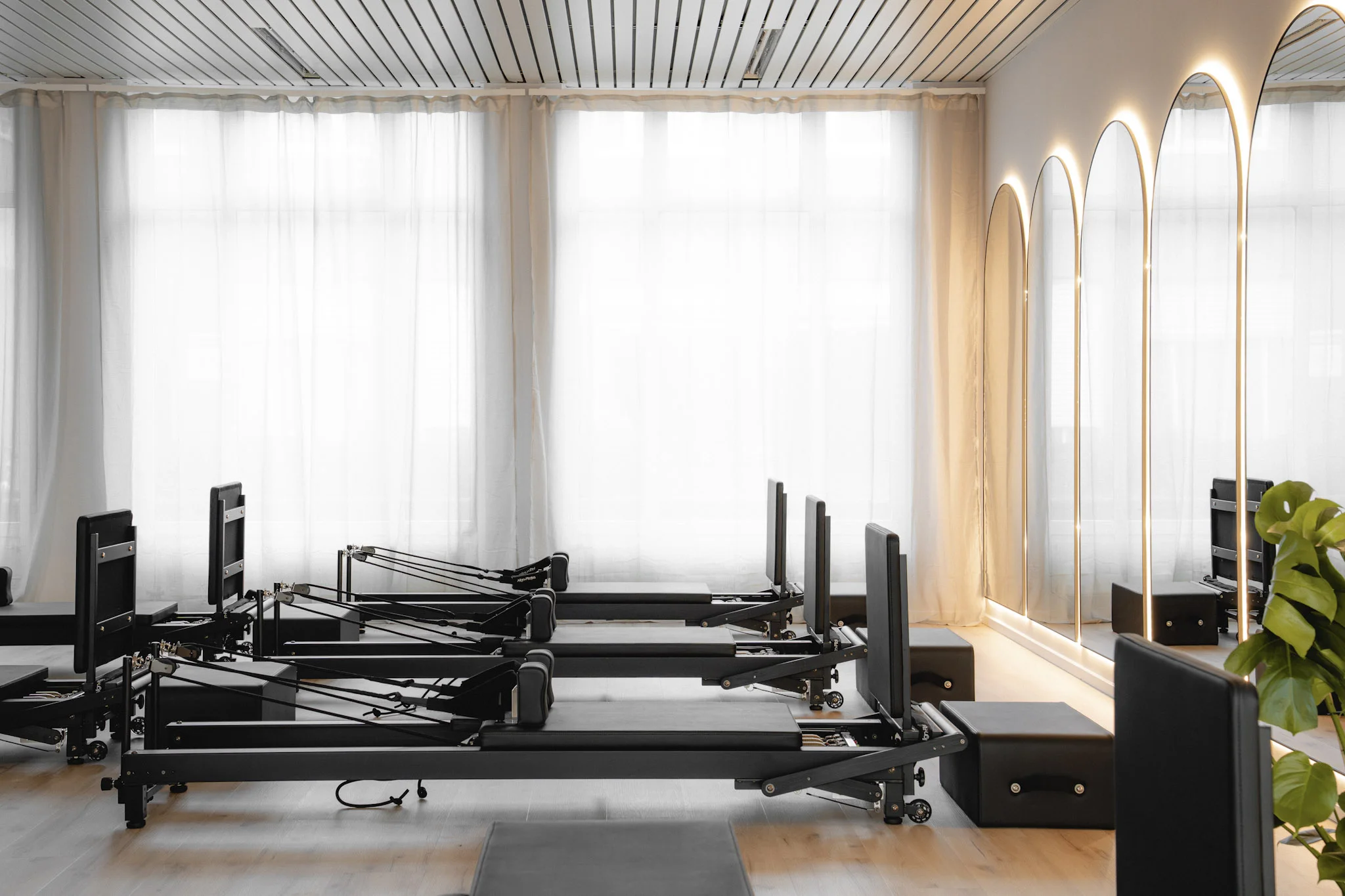 Heller Pilates-Raum mit drei schwarzen Reformer-Geräten vor großen weißen Vorhängen und vier hohen, beleuchteten Spiegeln.