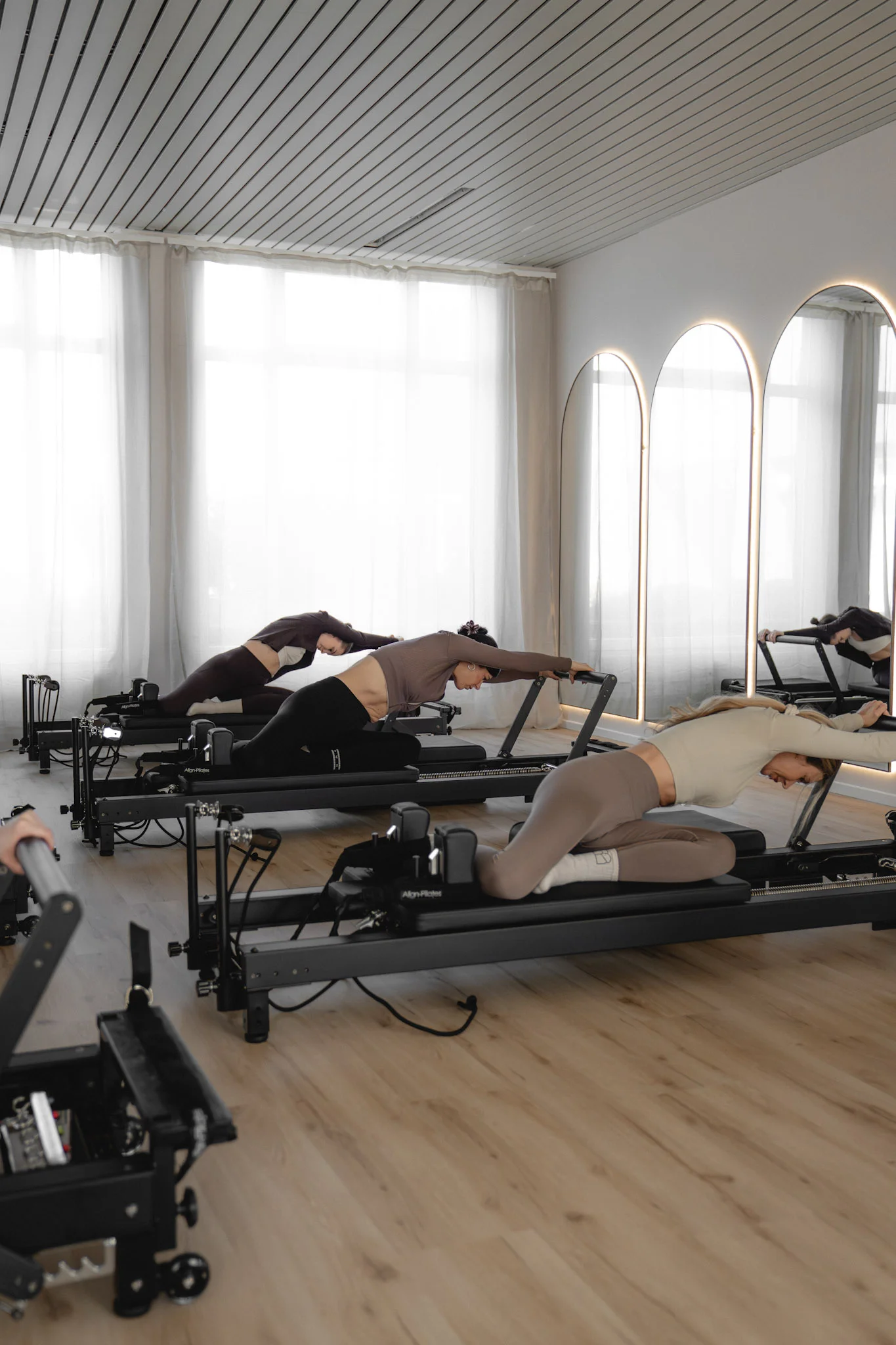 Drei Frauen im Fitnessstudio machen Pilates-Übungen auf Reformern vor großen Spiegeln.