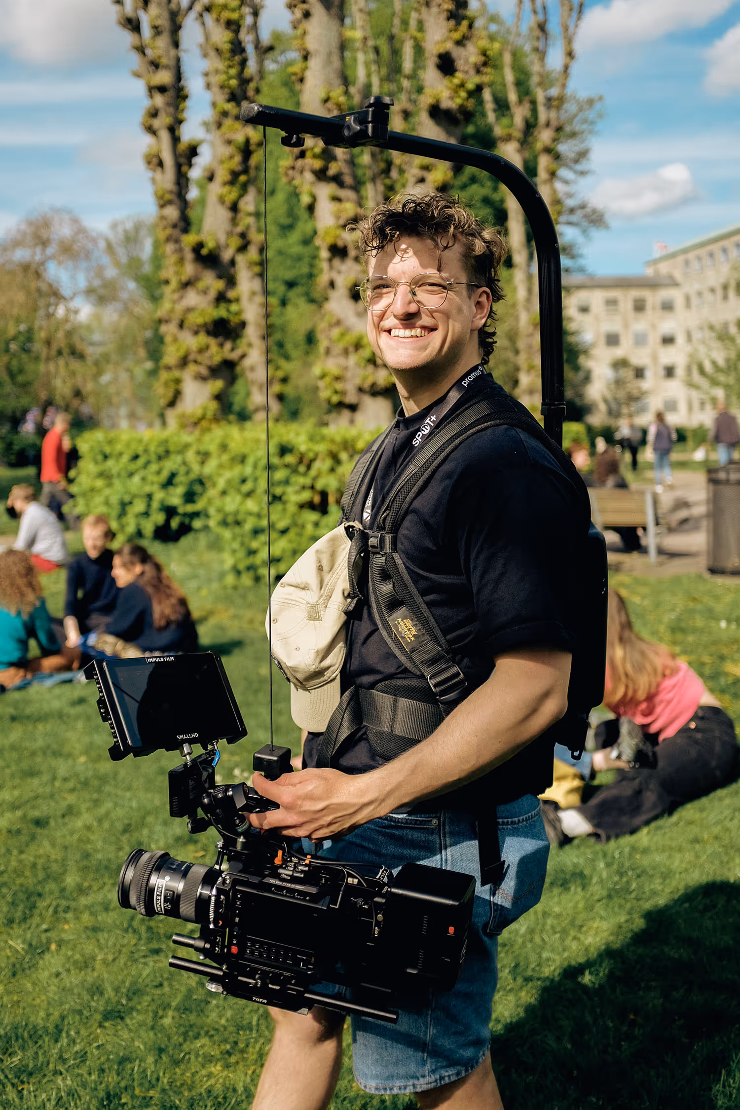 Smilende person med briller bærer et professionelt videokamera på en skuldermonteret stabilisator i en park.