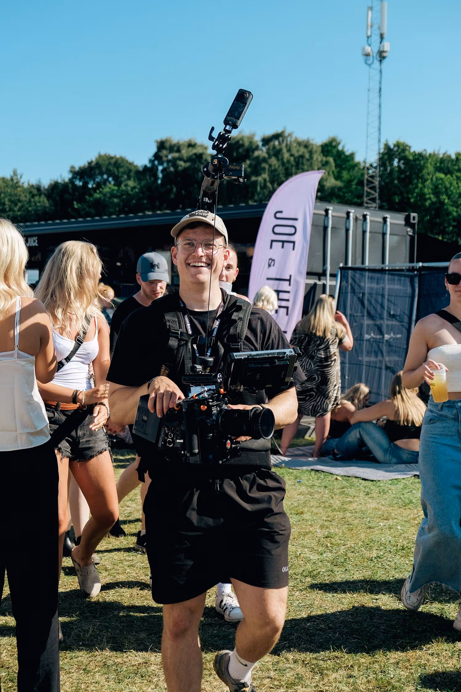 Mand med kasket og kameraudstyr smilende blandt folk ved udendørs festival.