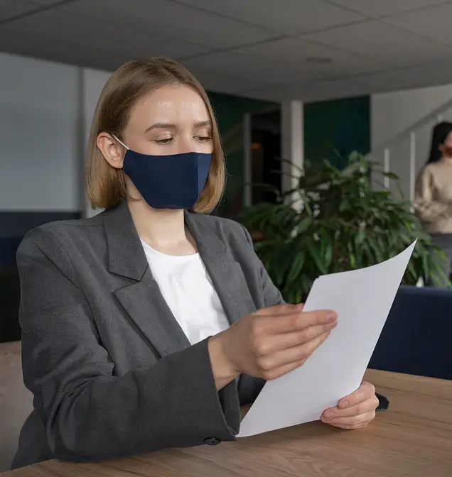 Woman wearing a navy face mask and gray blazer reading a document at a wooden desk in an office.