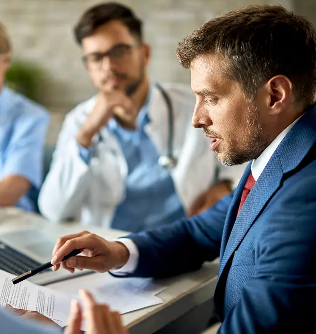 Man in blue suit pointing to a document while a doctor and other person listen attentively in a meeting.