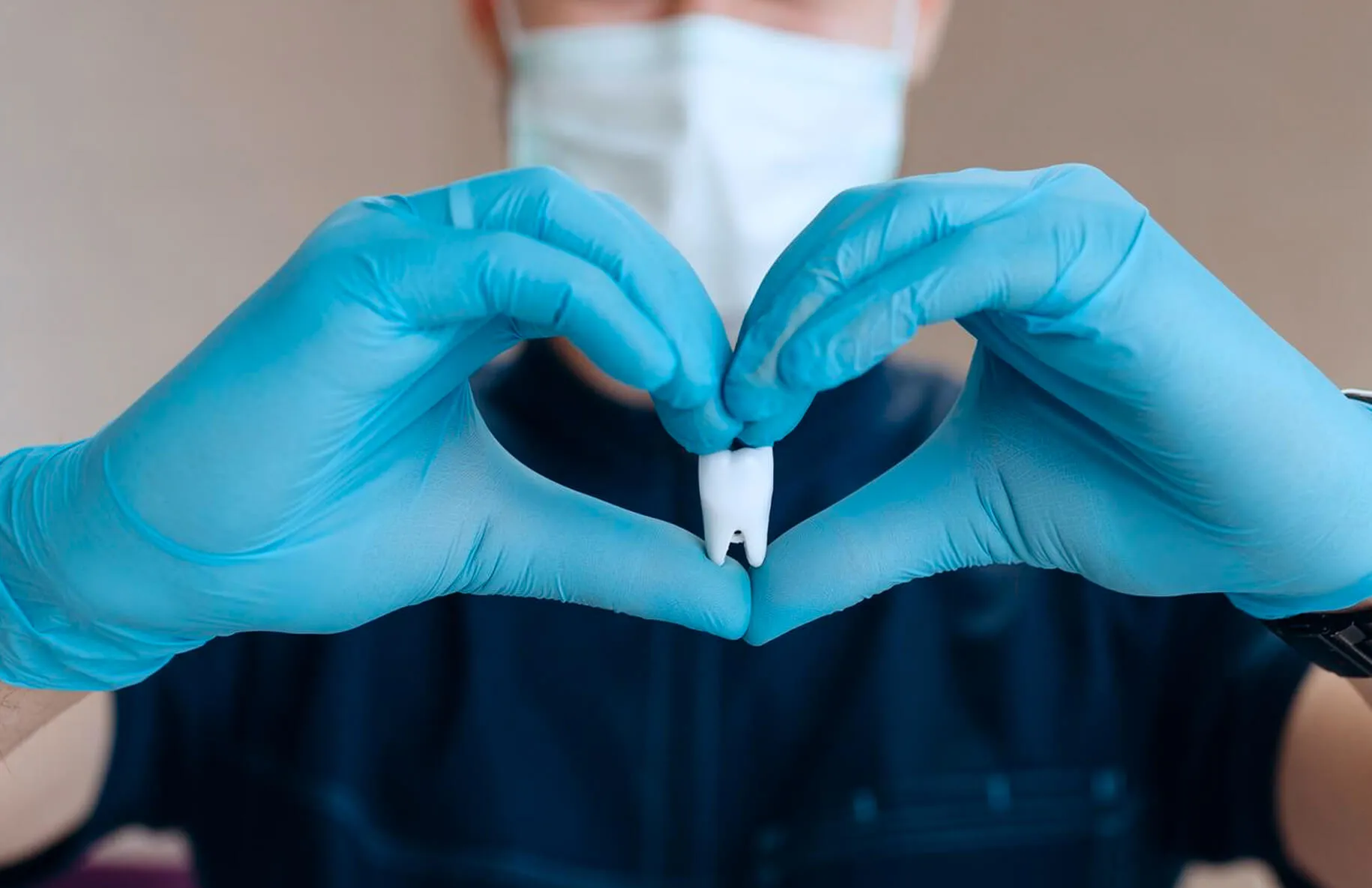 Person wearing blue gloves and a face mask forming a heart shape with hands around a model tooth.