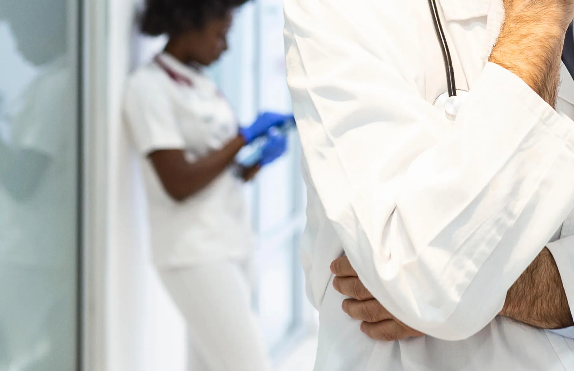 Close-up of a doctor in a white coat with a stethoscope, nursing staff blurred in the background.