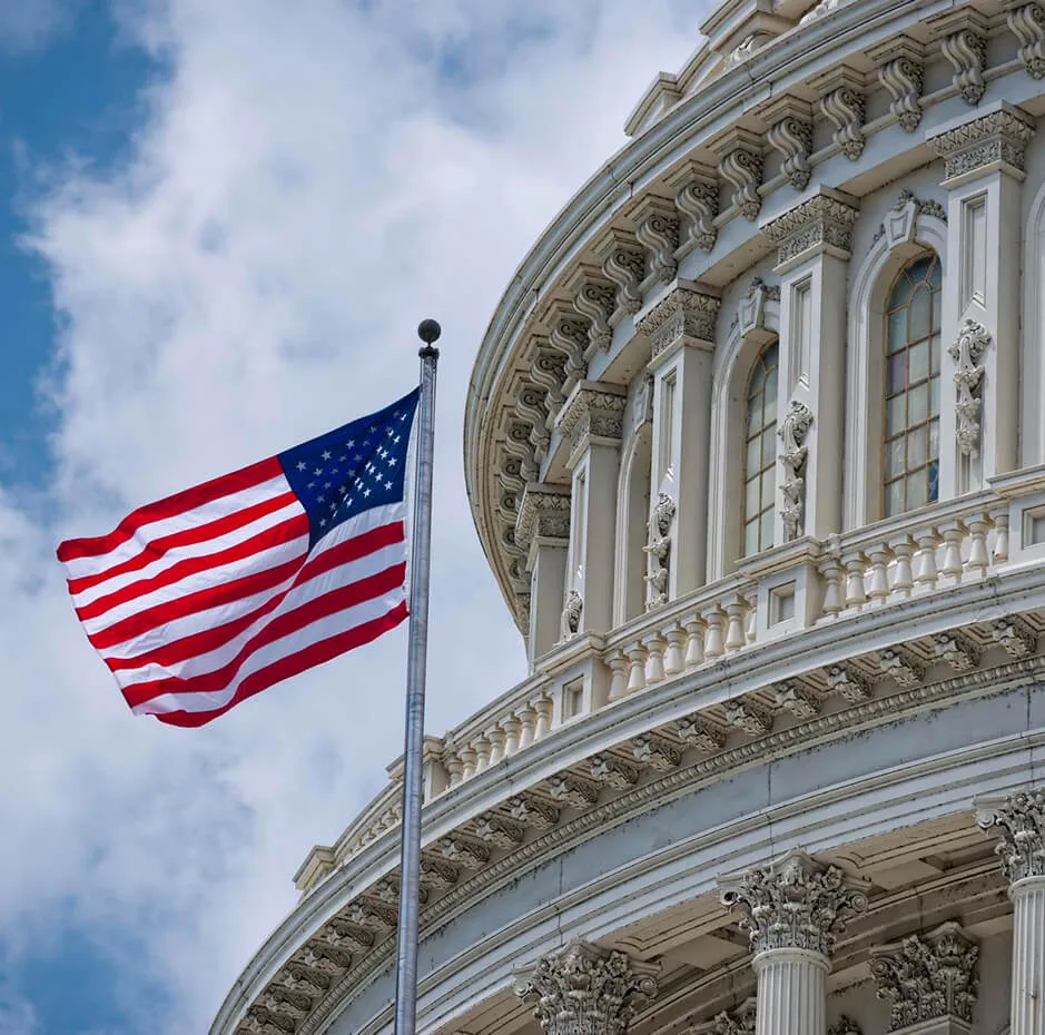 American flag waving on a flagpole beside the upper section of the United States Capitol building against a partly cloudy sky.
