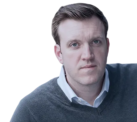 Portrait of a man with light skin, short brown hair, wearing a gray sweater over a white collared shirt, looking directly at the camera with a neutral expression.