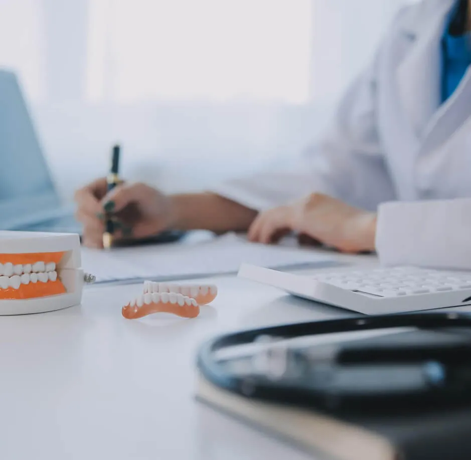 Dental models, a calculator, and a stethoscope on a desk with a person writing in the background.