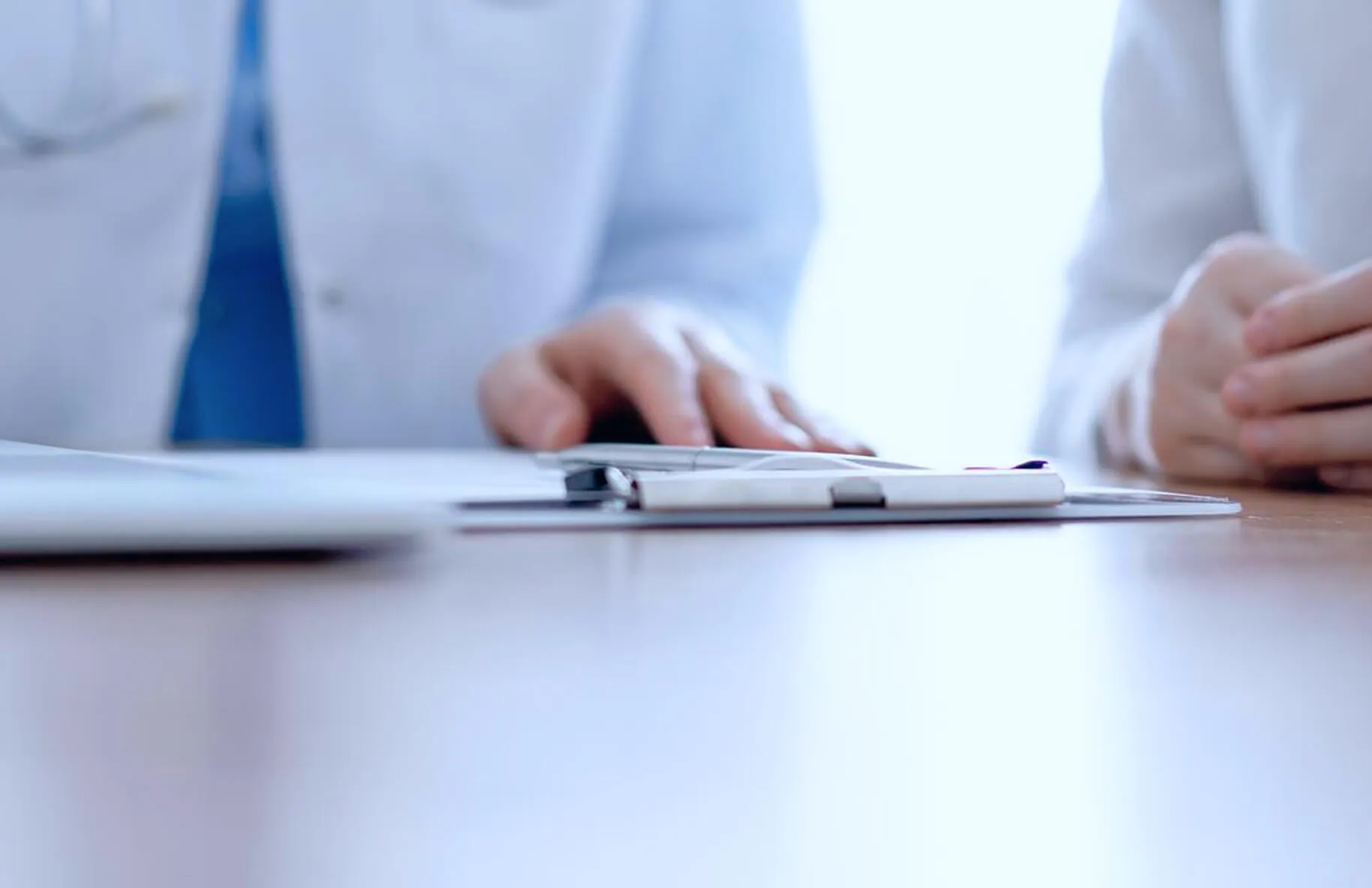 Close-up of hands and a clipboard on a wooden table with two people wearing white coats in the background.
