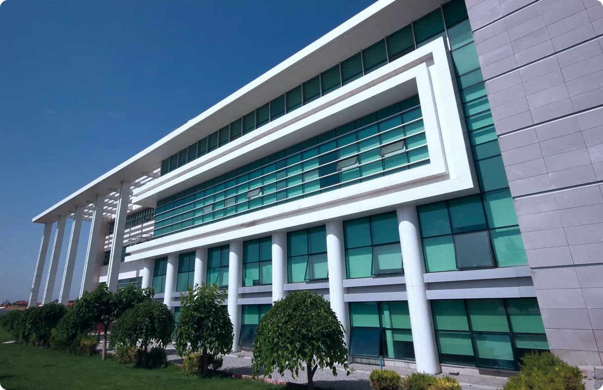 Modern office building with green tinted windows, white structural columns, and a landscaped lawn with bushes and small trees.