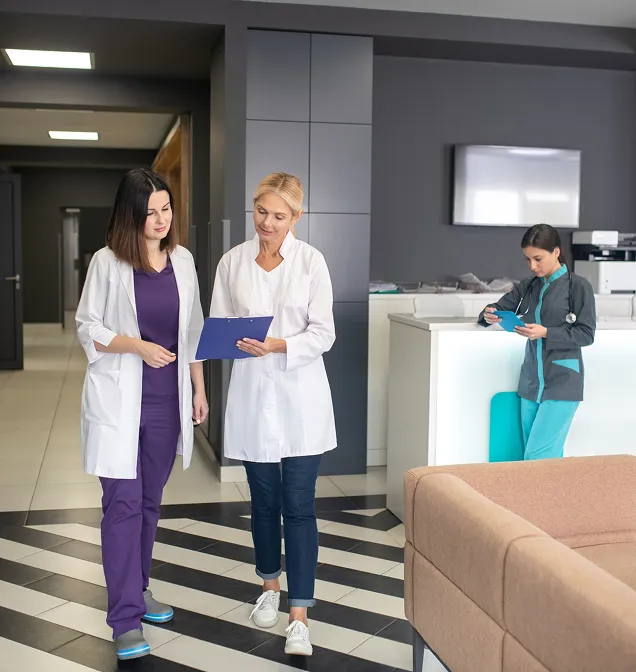Two female doctors walking and discussing notes on a clipboard in a modern medical facility while a third female healthcare worker checks a tablet near the reception desk.