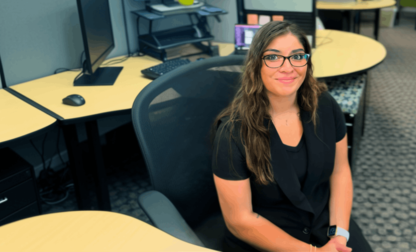 Smiling woman with glasses sitting in an office chair at a cubicle desk with computer monitors.