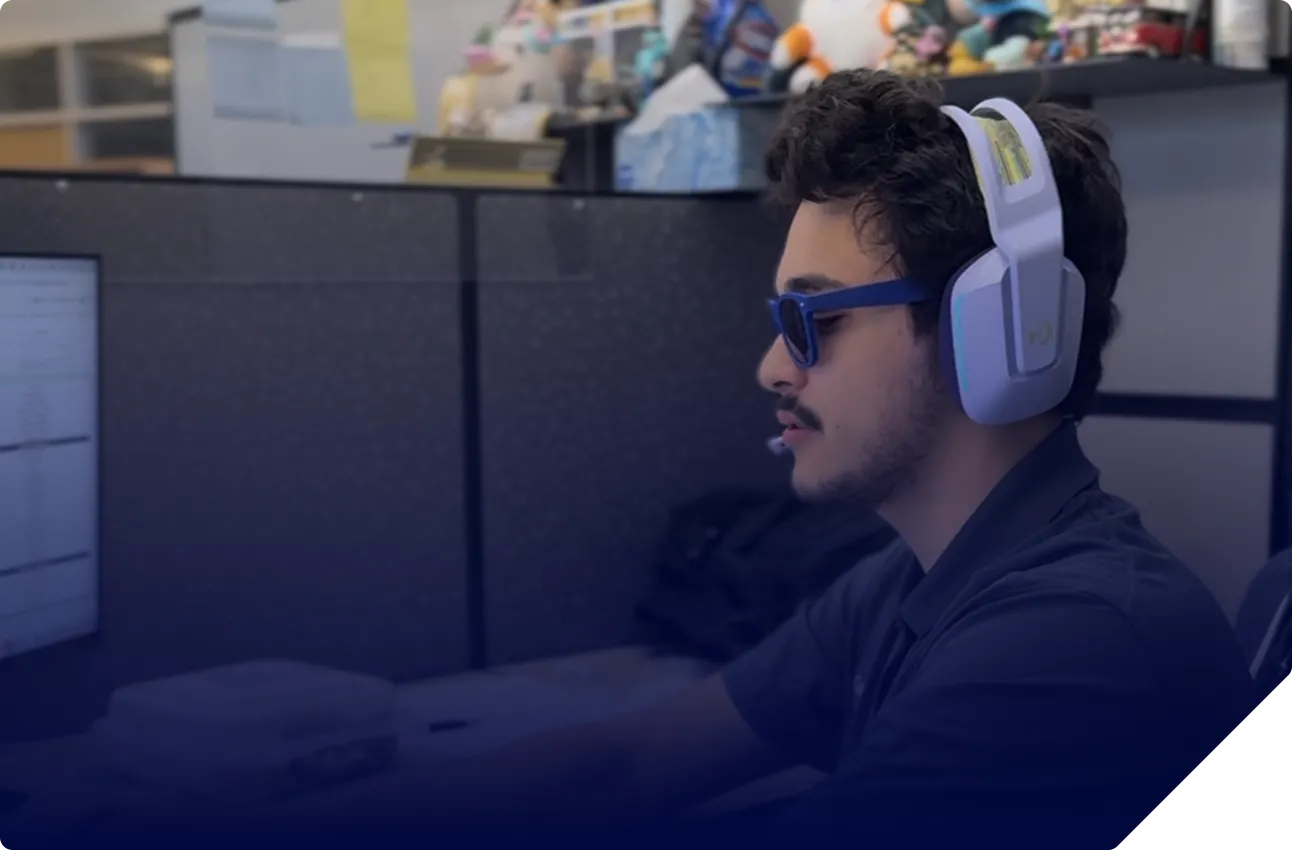 Man wearing blue sunglasses and white headset with microphone, working at a computer in an office cubicle.
