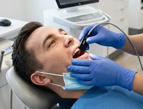 Dentist wearing blue gloves examining a male patient's mouth using a dental tool while the patient reclines in a dental chair.