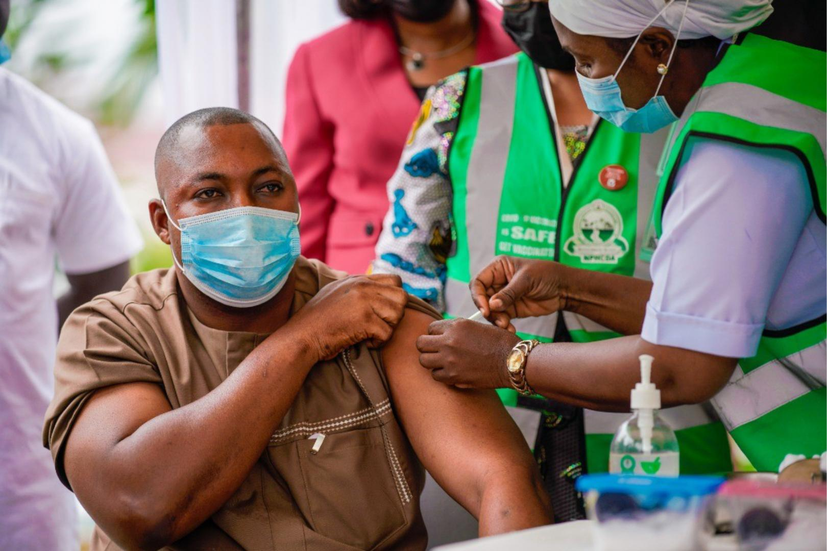 Man wearing a face mask receiving a vaccine injection from a healthcare worker in green safety vest and white uniform.