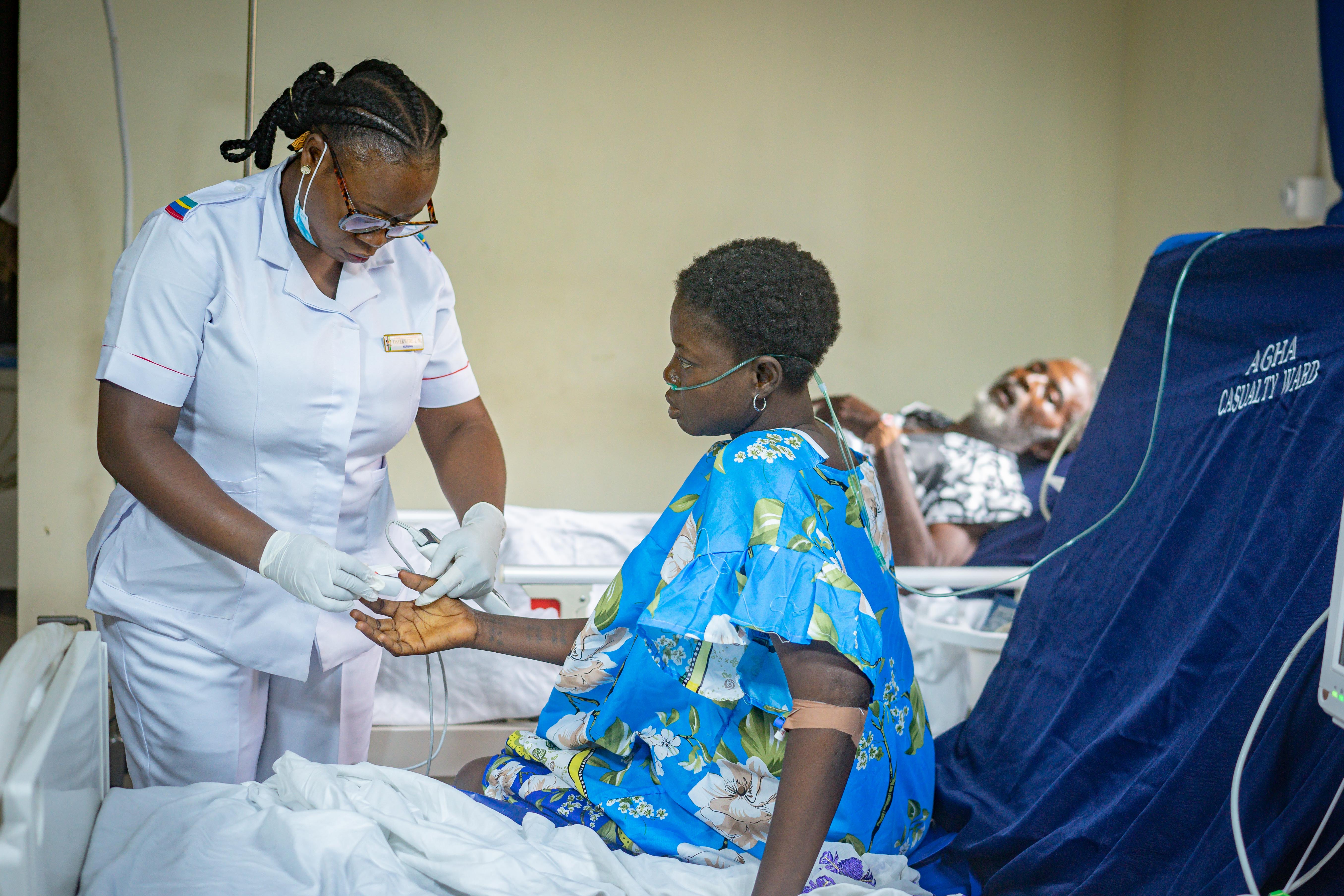 Nurse checking the pulse of a seated patient wearing a blue floral hospital gown with an oxygen tube, while another patient rests in a bed labeled 'Agha Casualty Ward.'