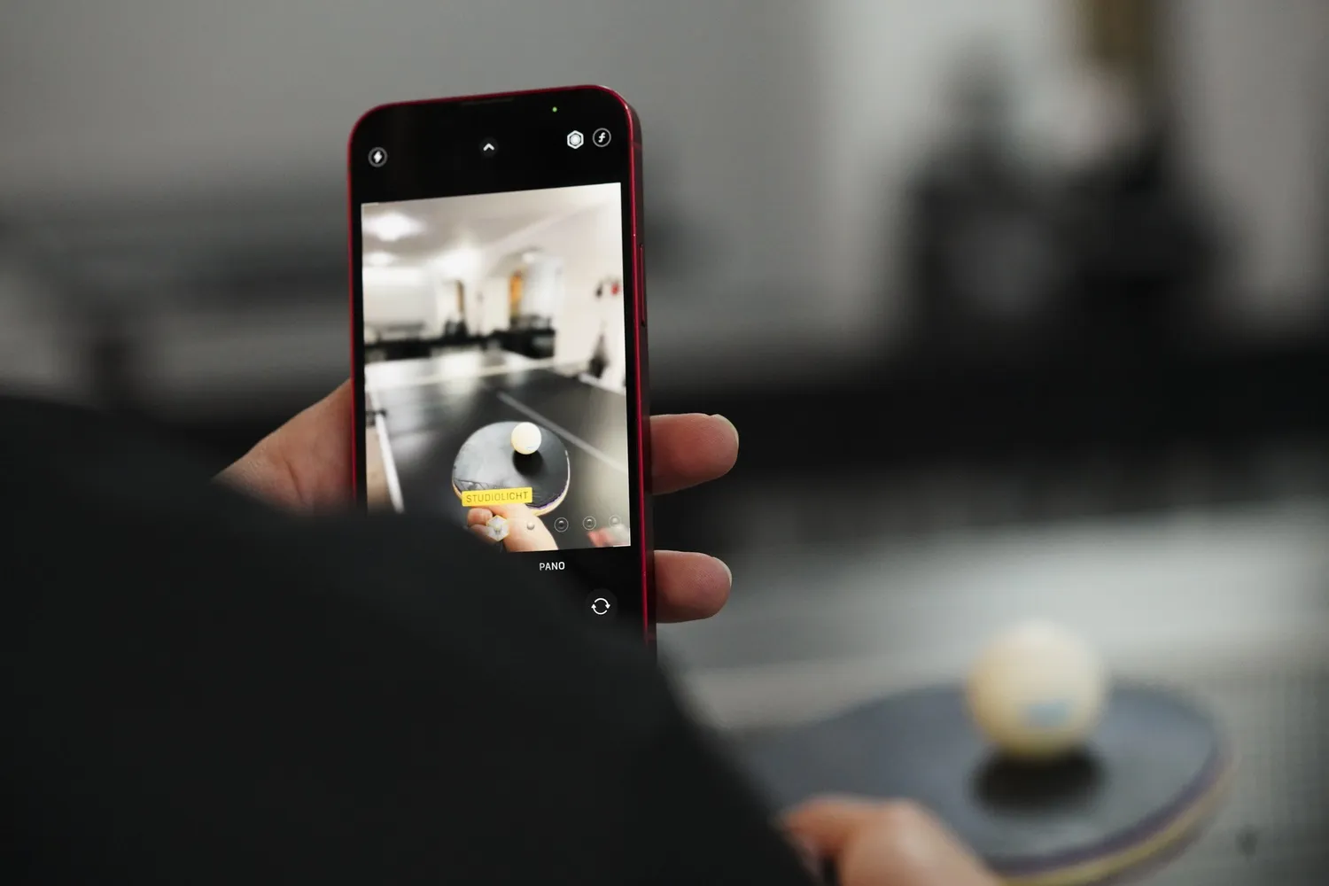 Hand hält Smartphone, das ein Tischtennisschläger mit Ball auf einem Tisch zeigt.