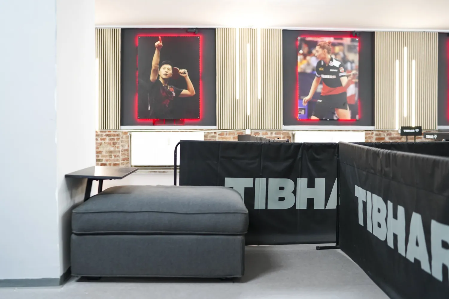 Grauer Sitzhocker neben Tisch, TIBHAR-Abtrennungen und beleuchtete Tischtennis-Spielerposter.