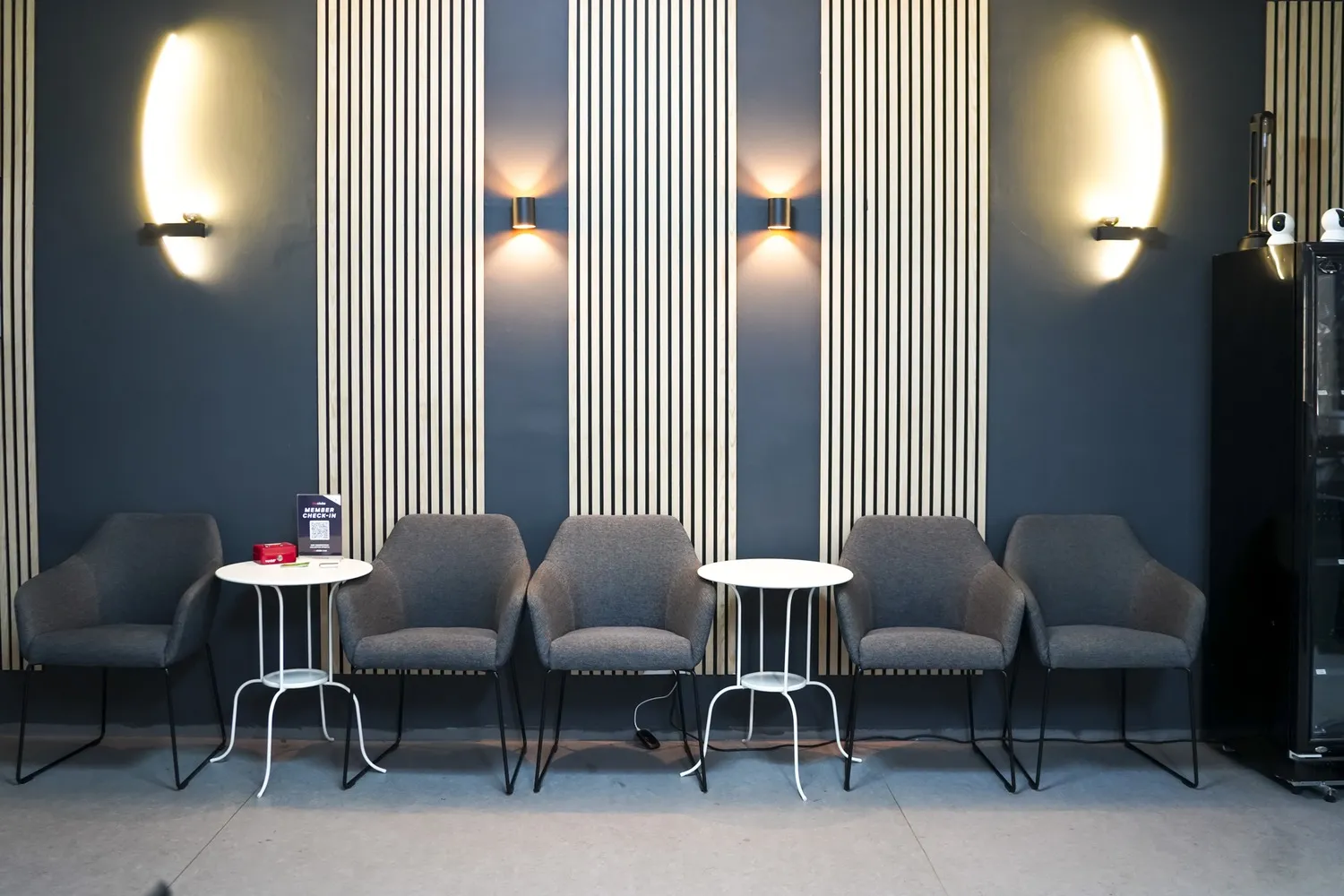 Fünf graue Stühle mit zwei kleinen weißen Tischen vor blau-grau gestreifter Wand in Wartebereich.