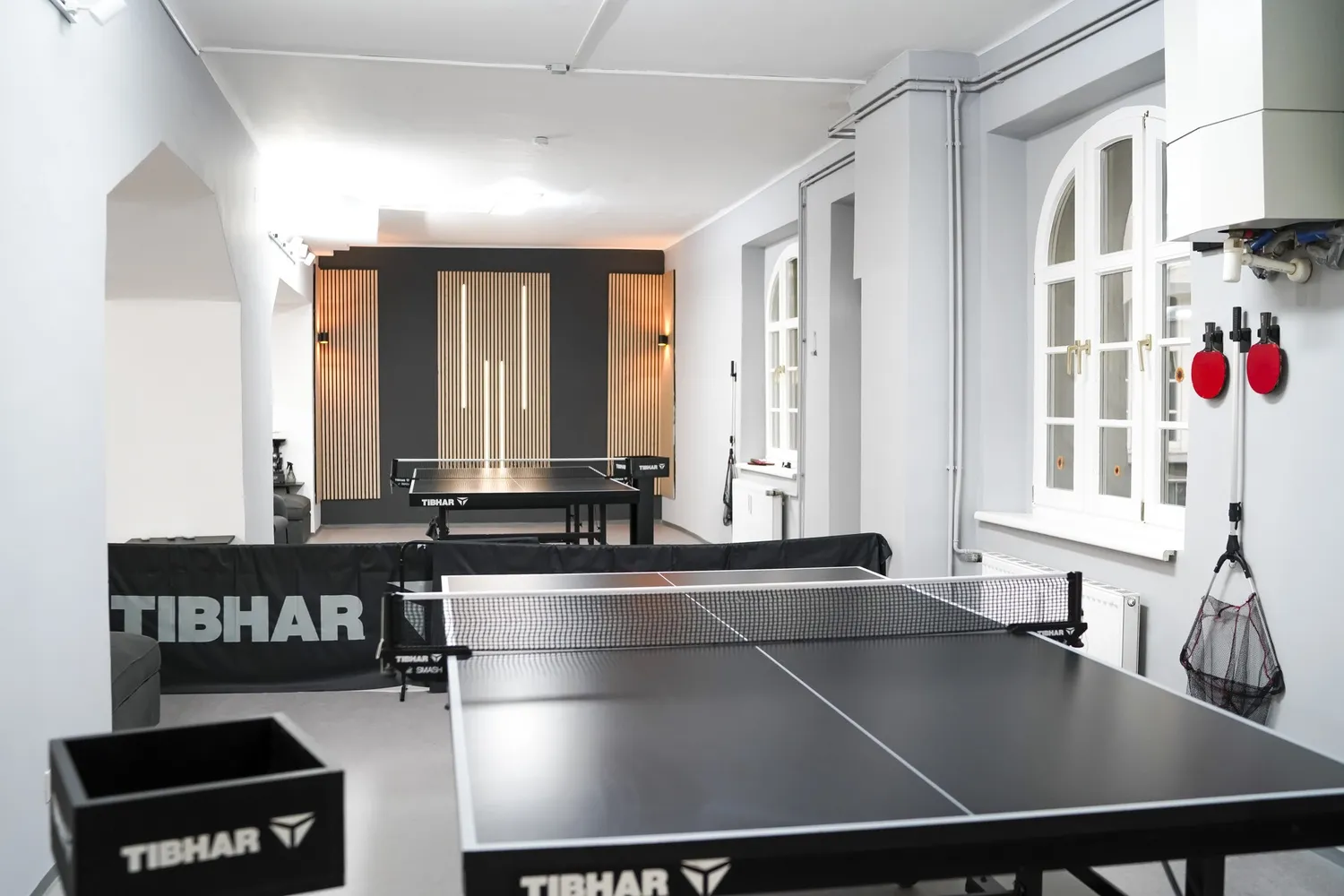 Innenraum mit zwei Tischtennisplatten, Wandlampen und Fenstern in einem hellen Raum.