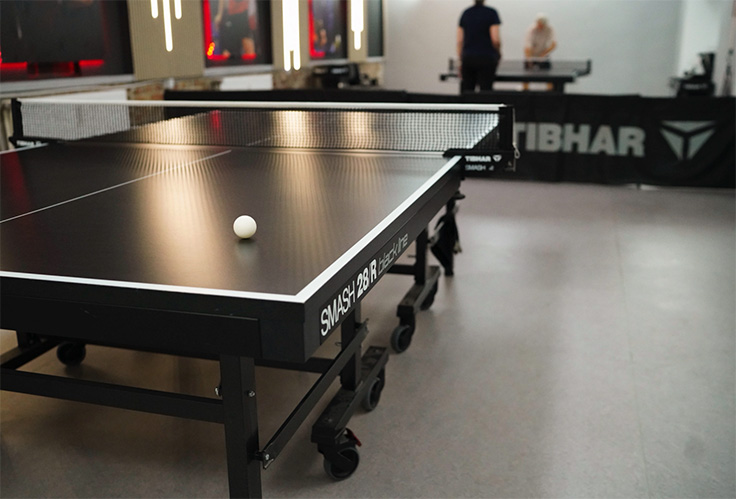 Schwarzer Tischtennistisch mit weißem Ball in einem Raum mit zwei spielenden Personen im Hintergrund.