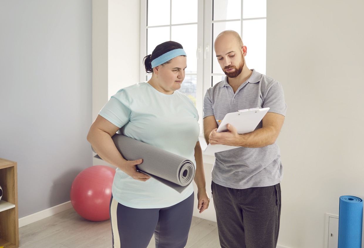 Fitness coach reviewing notes on clipboard with woman holding a yoga mat in a bright exercise room.