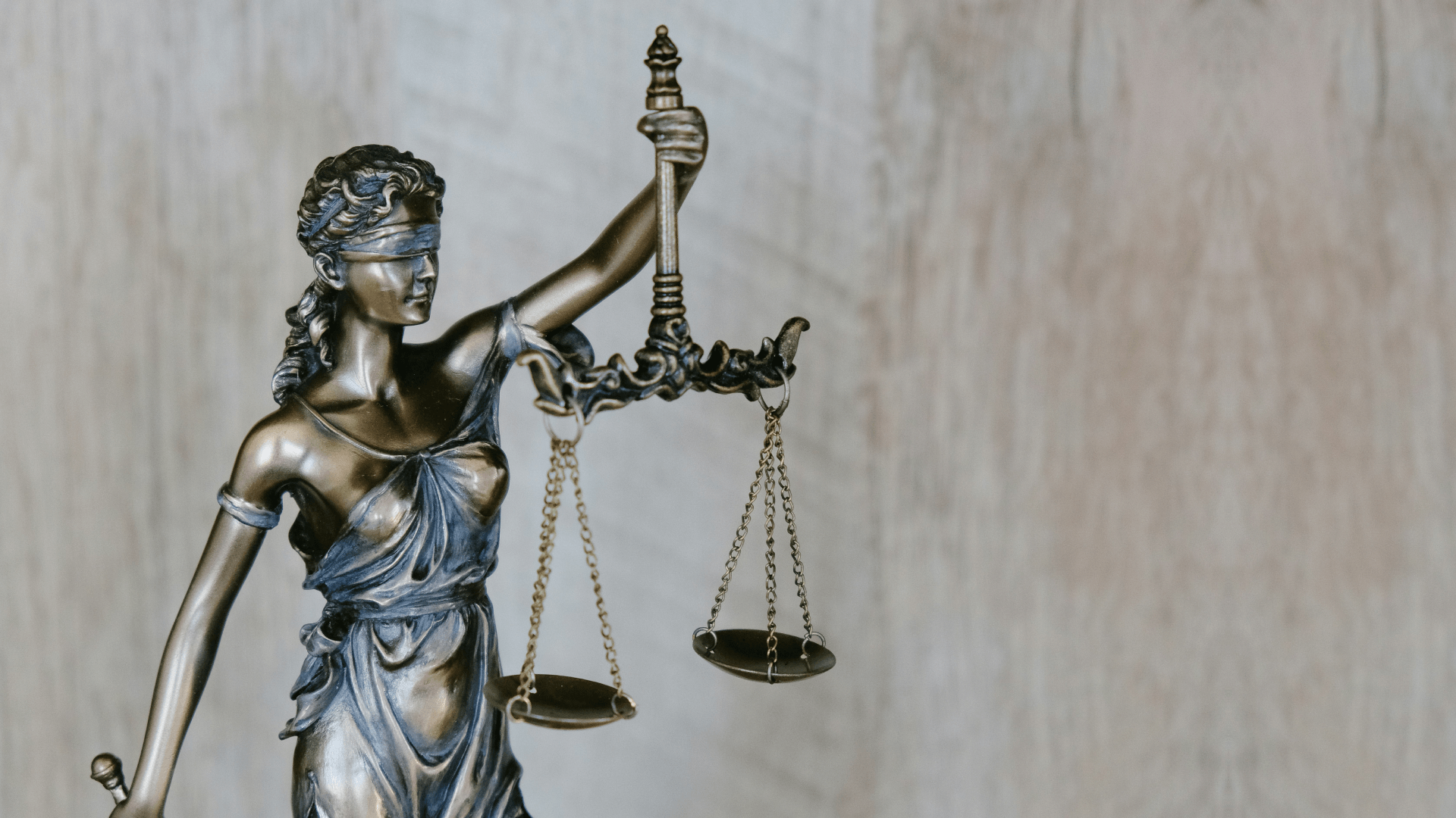Statue der Justitia mit verbundenen Augen und Waage vor unscharfem hellbraunem Hintergrund.