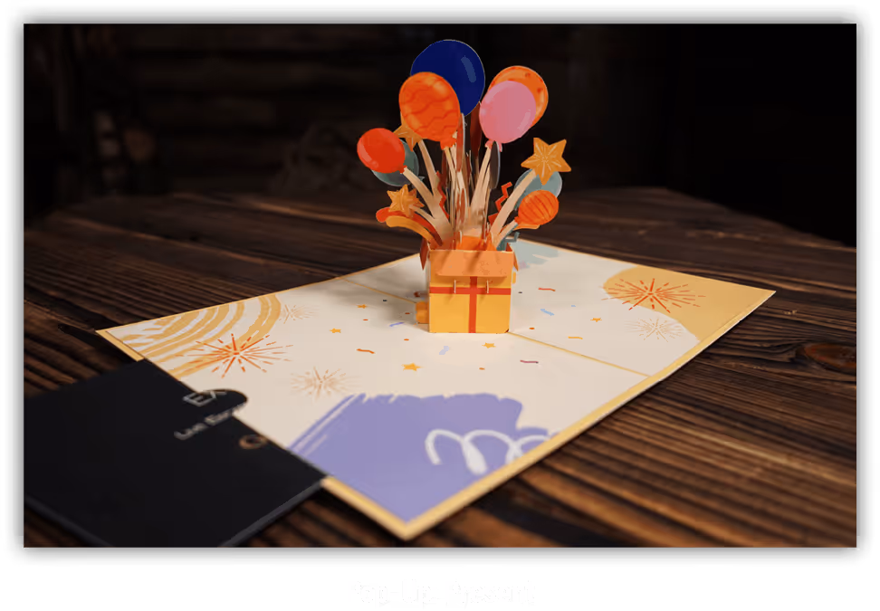 Pop-up-Grußkarte für einen Escape Room mit einem geöffneten gelben Geschenk und bunten Luftballons, Sternen und Feuerwerksmotiven auf einem Holztisch.