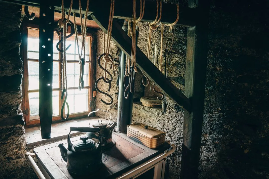 Old rustic kitchen scene with iron kettles on a wooden stove and various metal hooks hanging from ropes near a window.