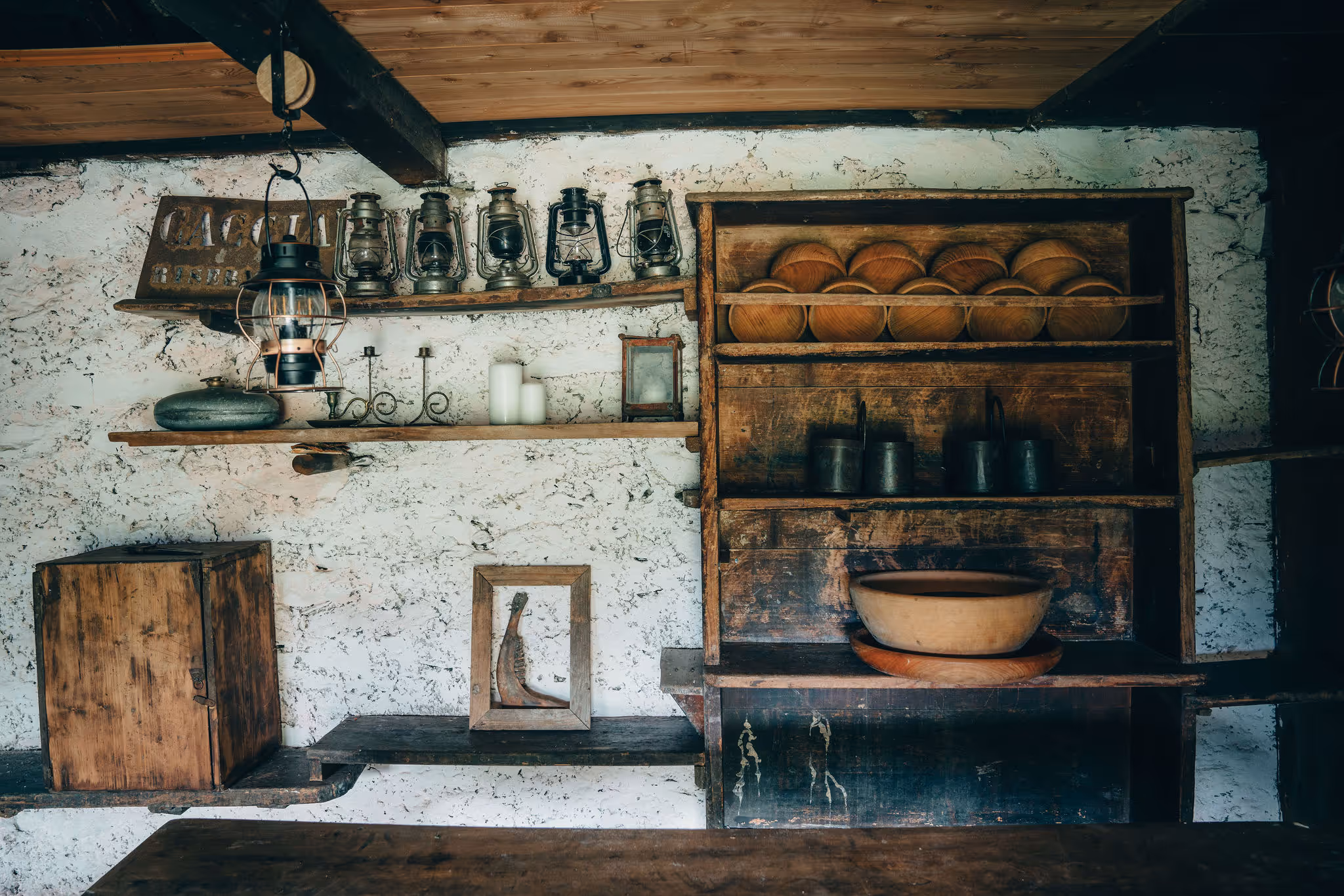 Rustic interior wall with wooden shelves holding old lanterns, candles, pots, and bowls, showcasing vintage kitchenware.
