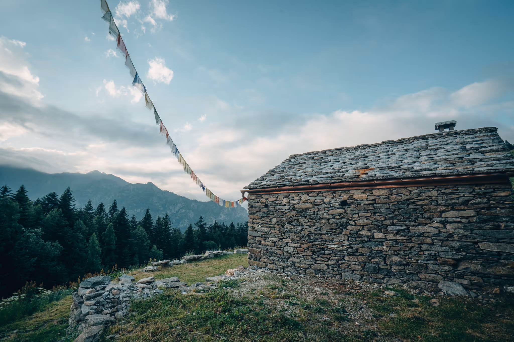 Stone cabin with a slate roof in a mountainous landscape during cloudy dusk, with trees and a mountain range in the background and colorful prayer flags strung from the cabin.