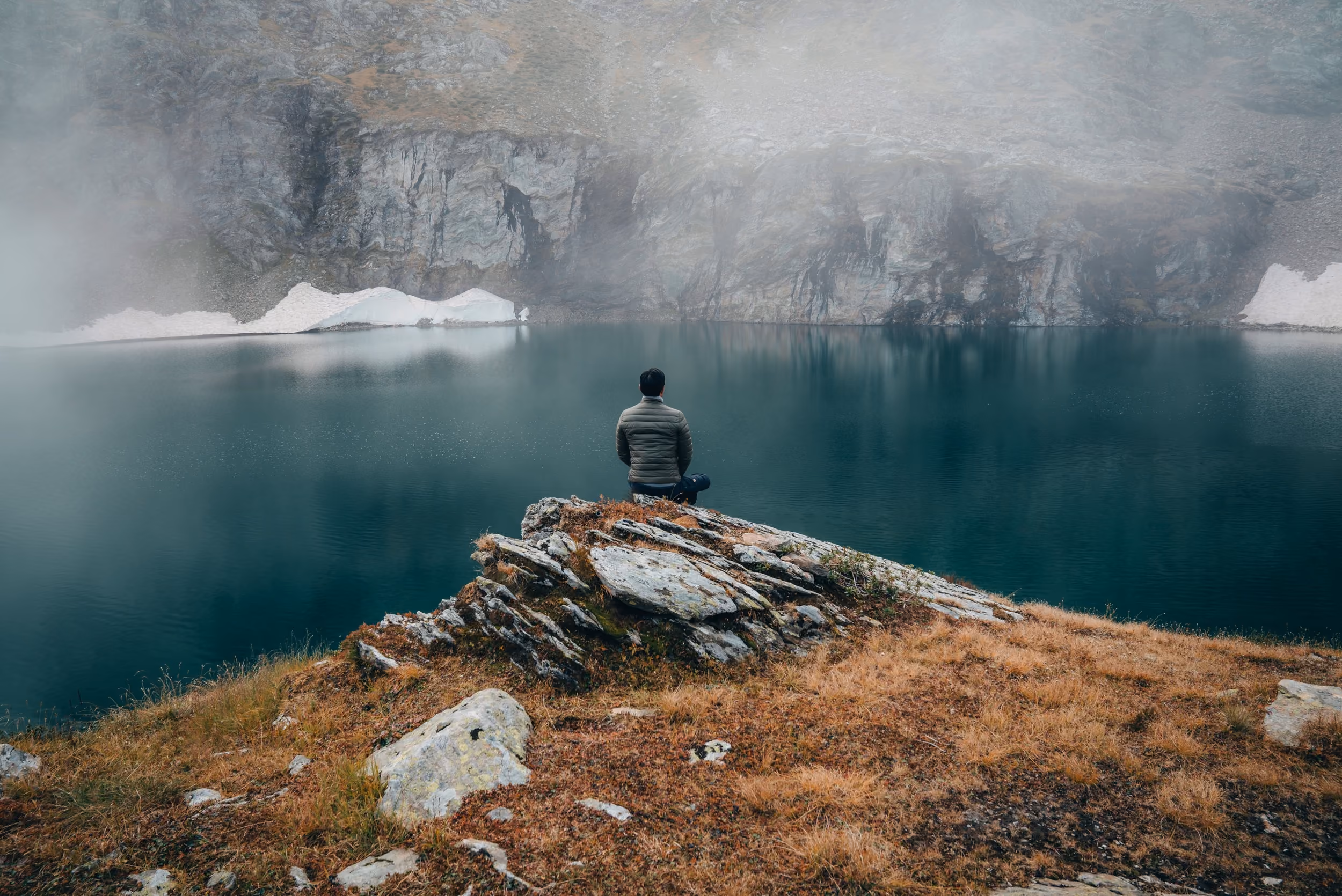 A person sitting cross-legged on a rocky outcrop facing a misty mountain lake surrounded by steep cliffs with patches of snow.