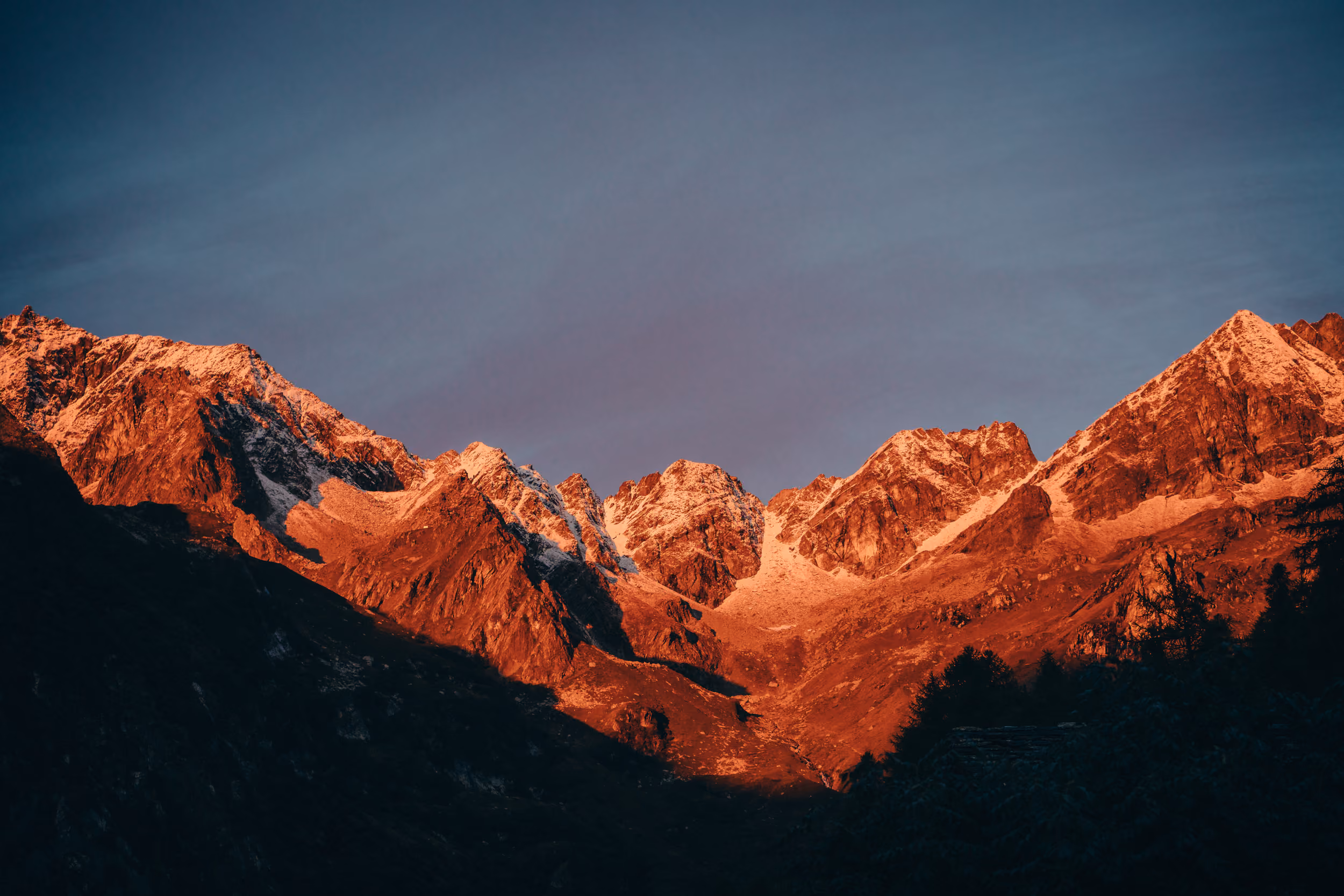 Mountain range with snow-capped peaks illuminated by warm sunlight against a clear sky at dusk or dawn.