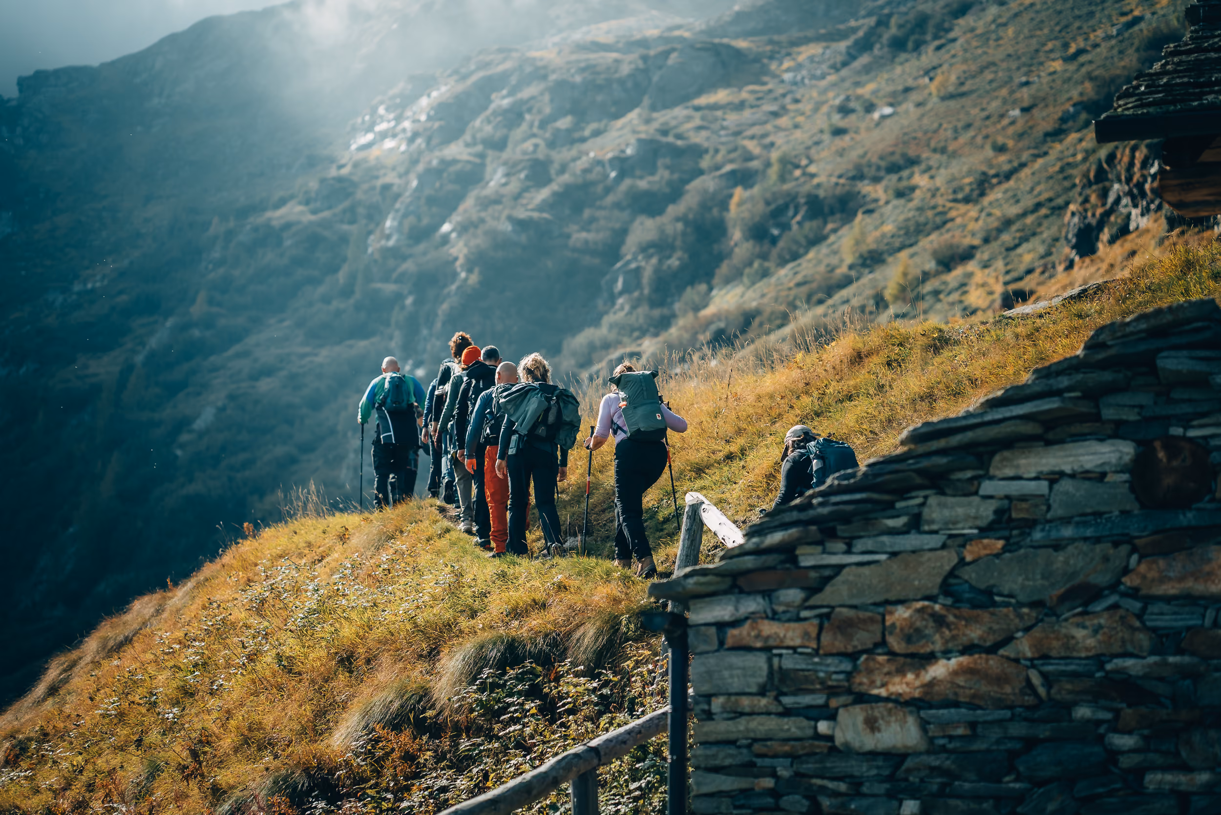 A group of hikers walking in single file along a grassy mountain trail beside a stone structure, with wooded hills in the background and sunlight breaking through the mist.