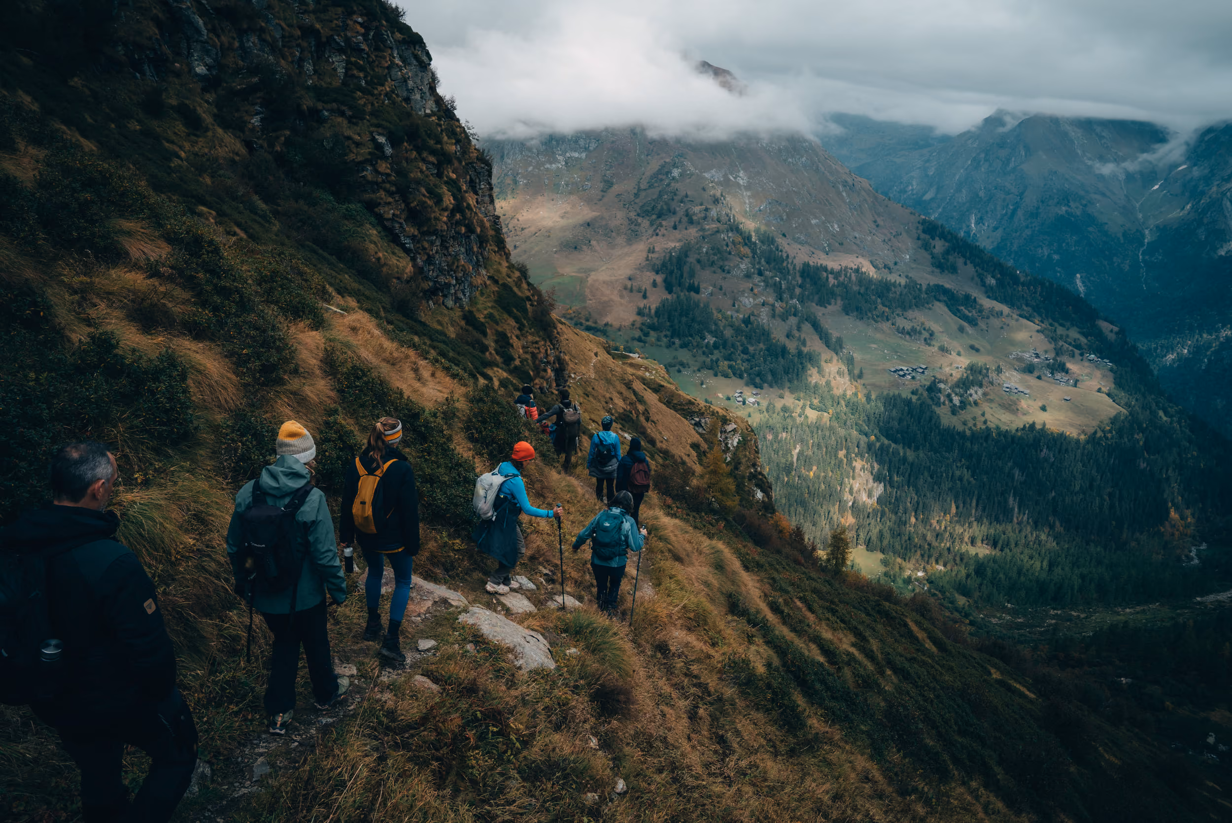 Group of hikers walking along a narrow mountain trail on a grassy slope with steep rocky cliffs on the left and expansive forested valleys and mountains in the distance under a cloudy sky.
