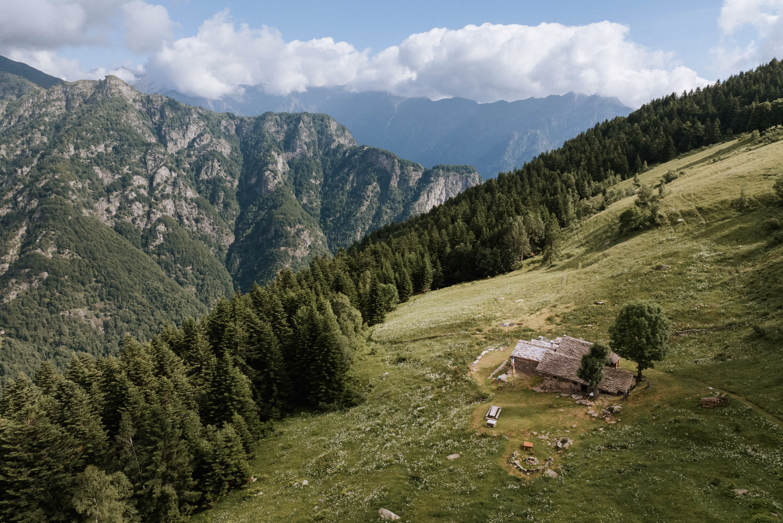 Aerial view of a rustic stone cabin with a tiled roof on a grassy hillside surrounded by dense evergreen forest, with rugged mountain peaks and a cloudy sky in the background.
