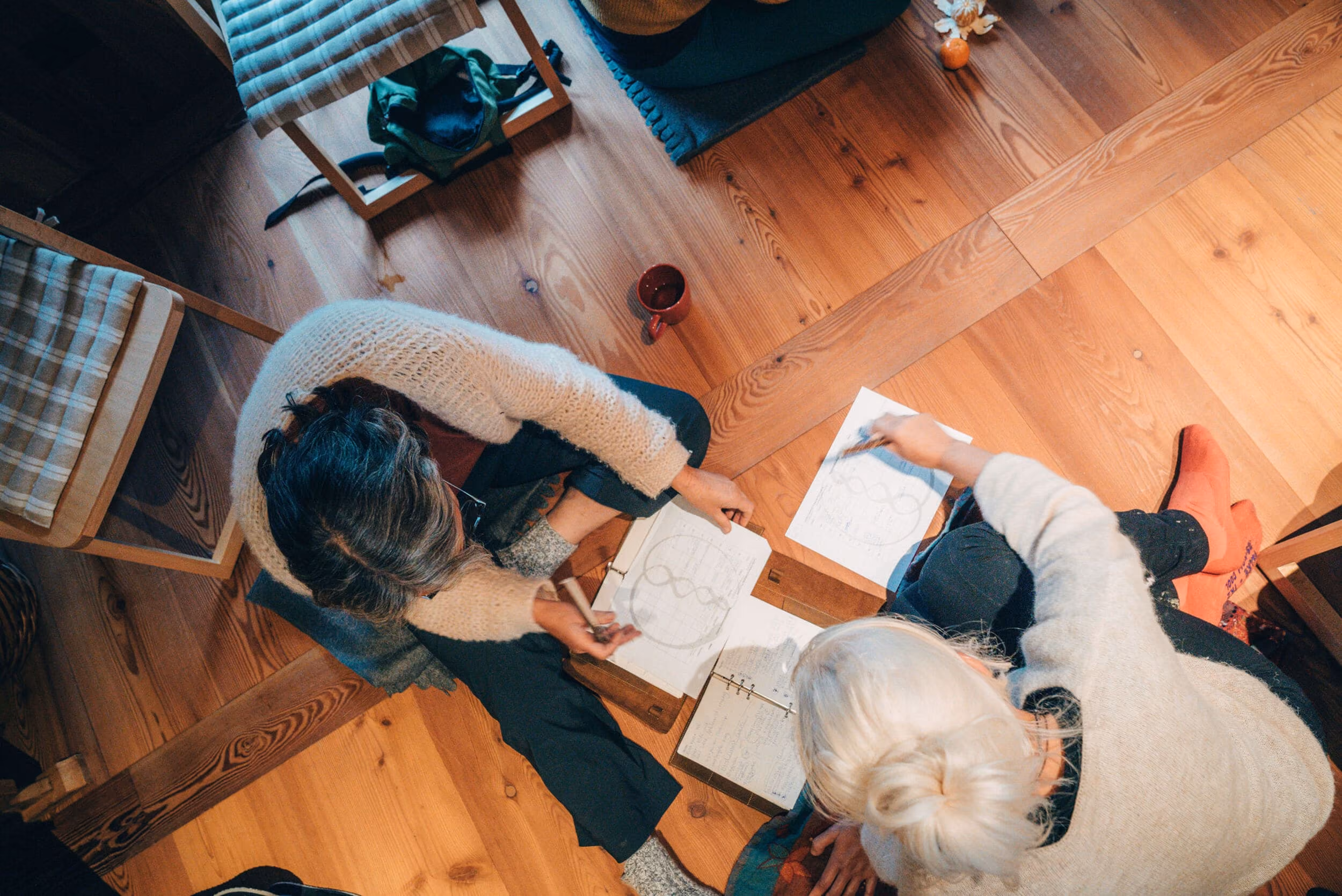 Top view of two people sitting on a wooden floor drawing on papers with pencils, surrounded by chairs and a cup.