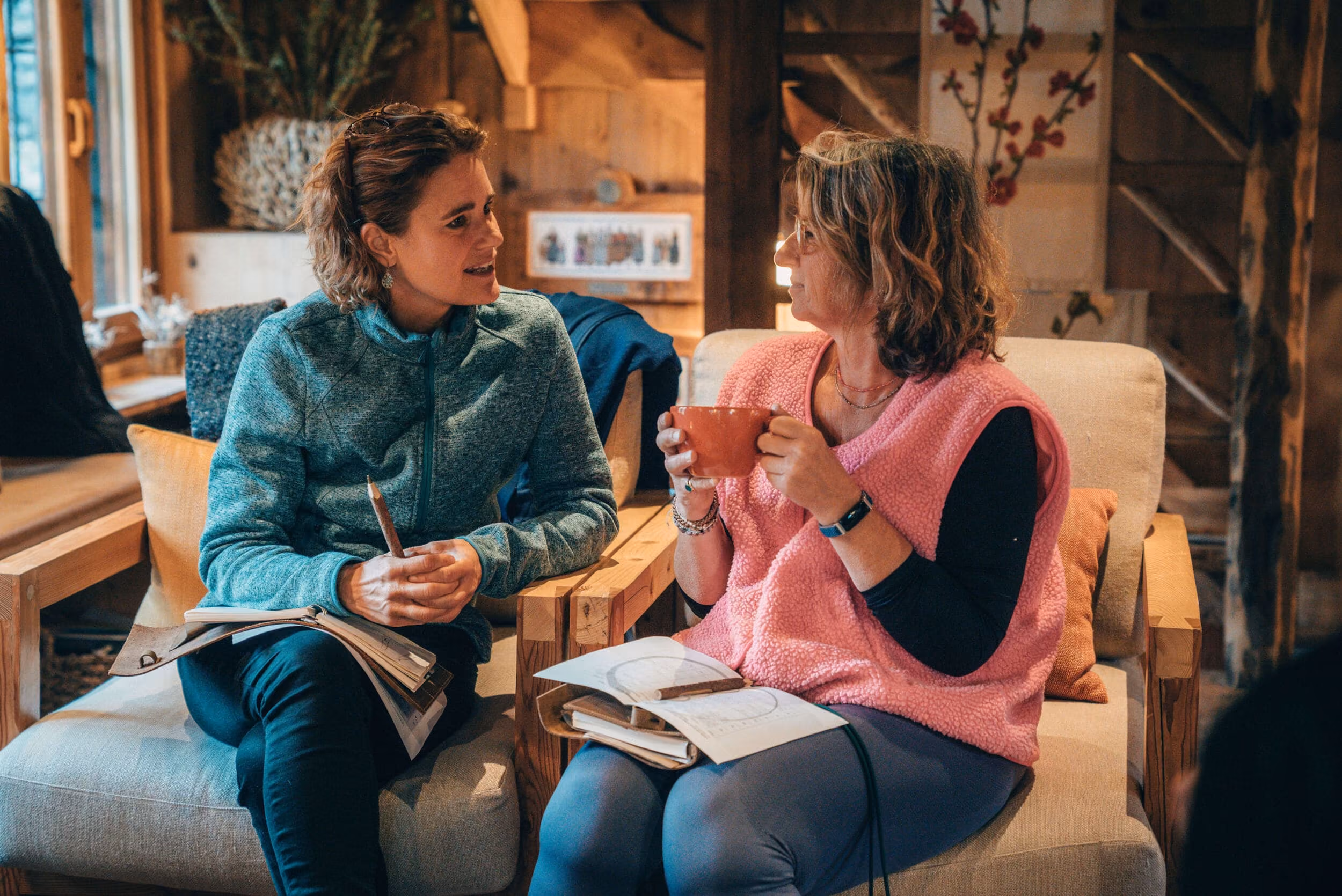 Two women sitting on cushioned armchairs in a cozy wooden room, engaged in conversation; one holds a pencil and notebook while the other holds an orange mug with notebooks on her lap.