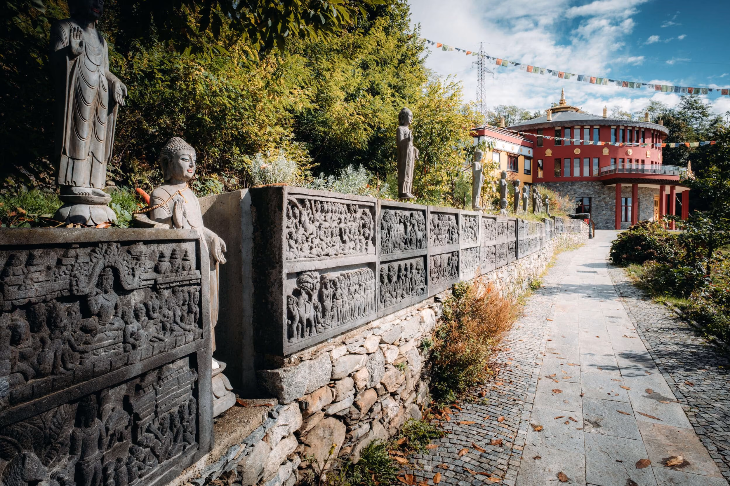 Outdoor pathway beside a stone wall with detailed Buddhist carvings and standing Buddha statues, leading to a round building adorned with prayer flags under a partly cloudy sky.