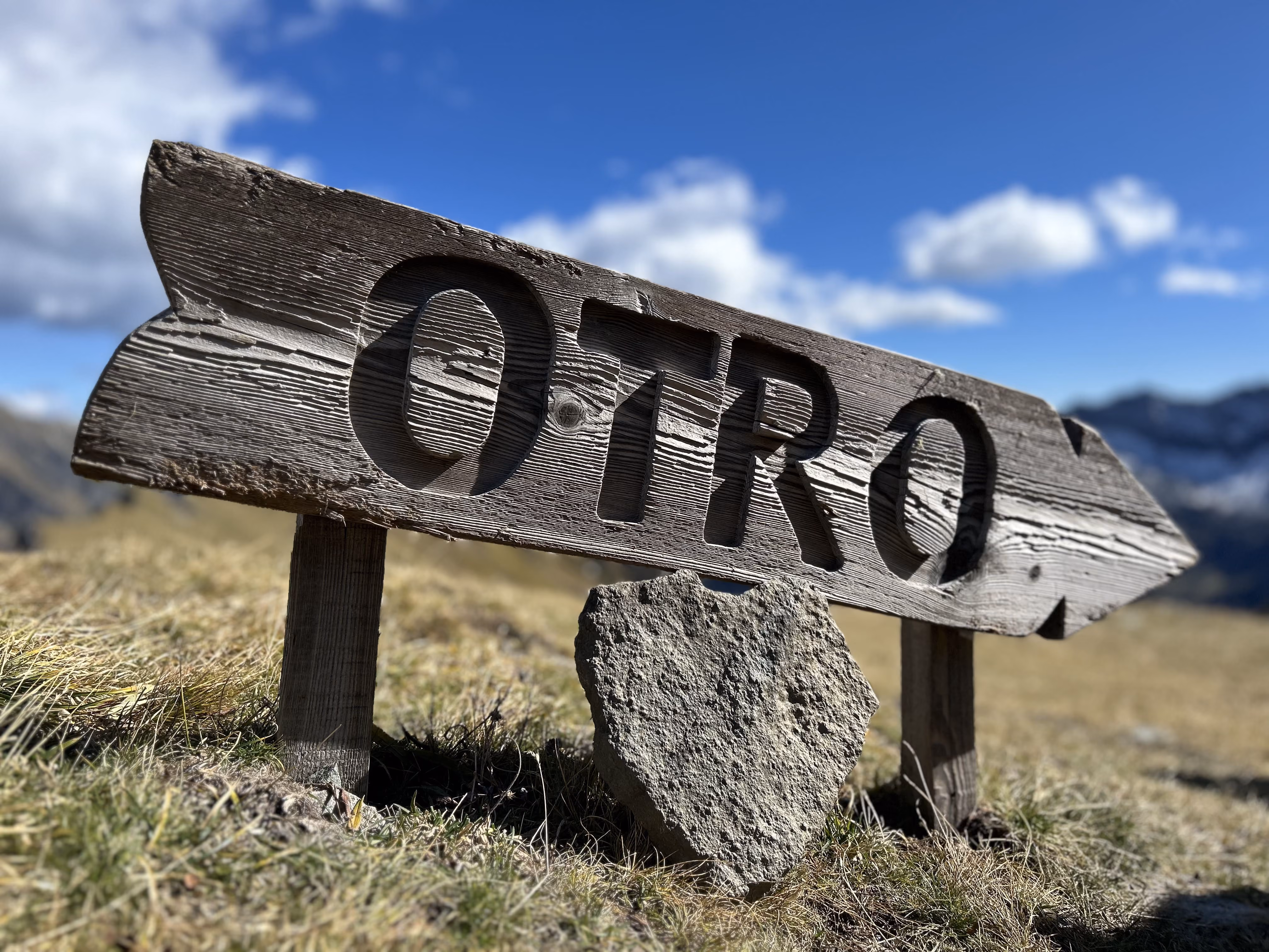 Weathered wooden sign with the word OTRO carved into it, placed on grass with a stone shield at its base, under a blue sky with clouds and mountains in the background.