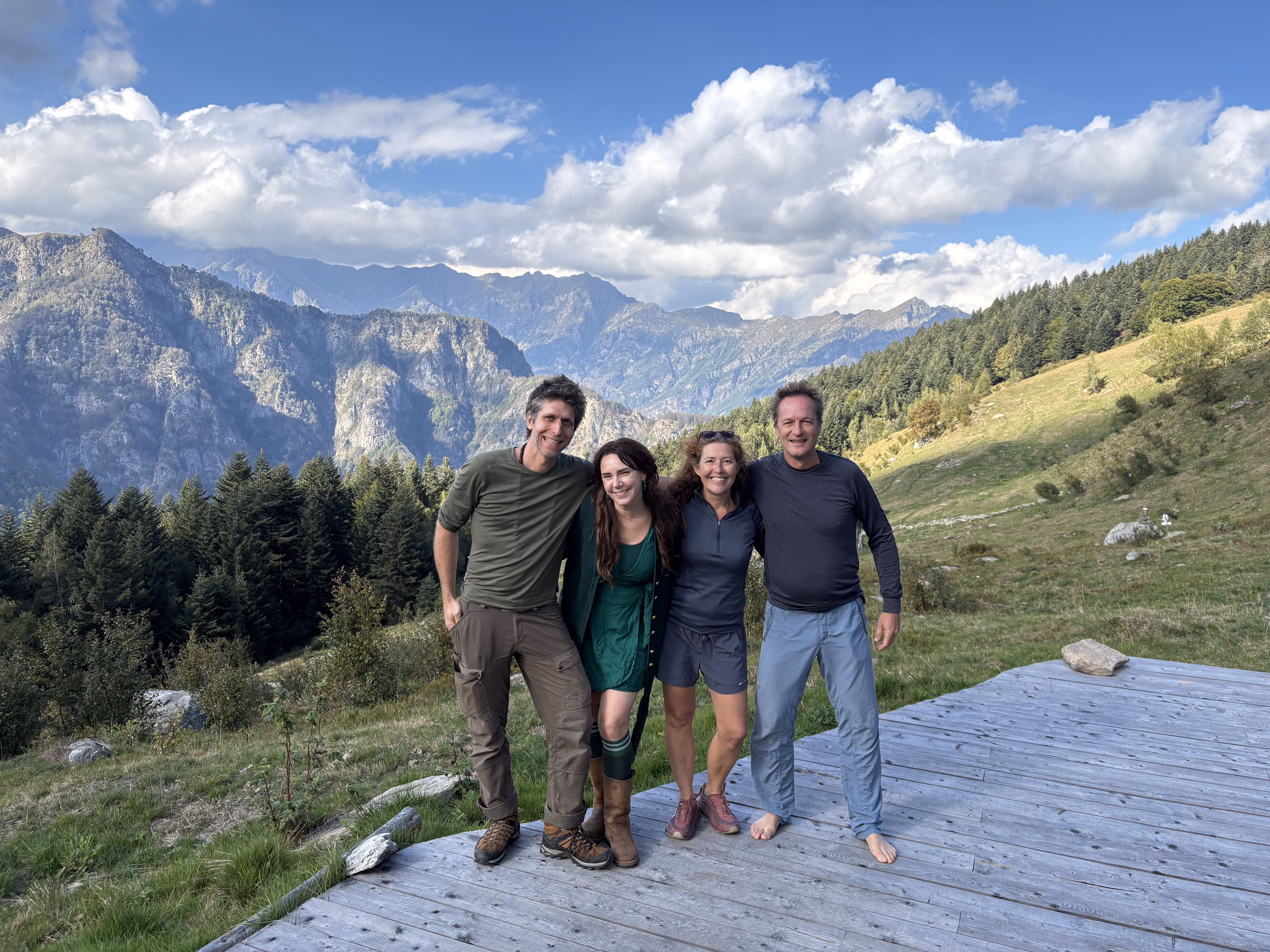 Four adults standing close together on a wooden deck in a mountainous outdoor setting with green hills, pine trees, and partly cloudy blue sky in the background, two men and two women smiling at the camera, one man barefoot.