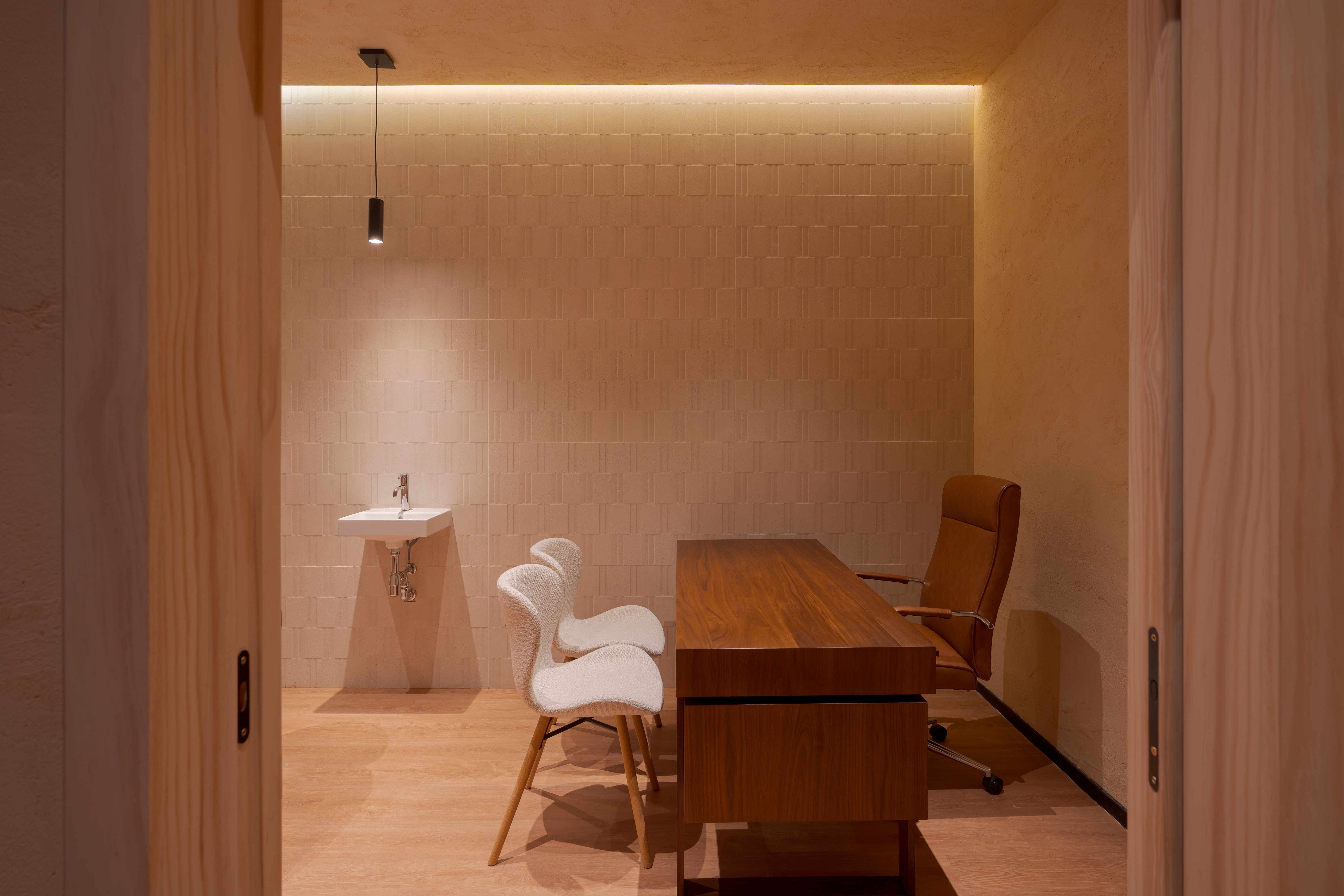 Oficina minimalista con escritorio de madera, tres sillas y lavabo en pared texturizada.
