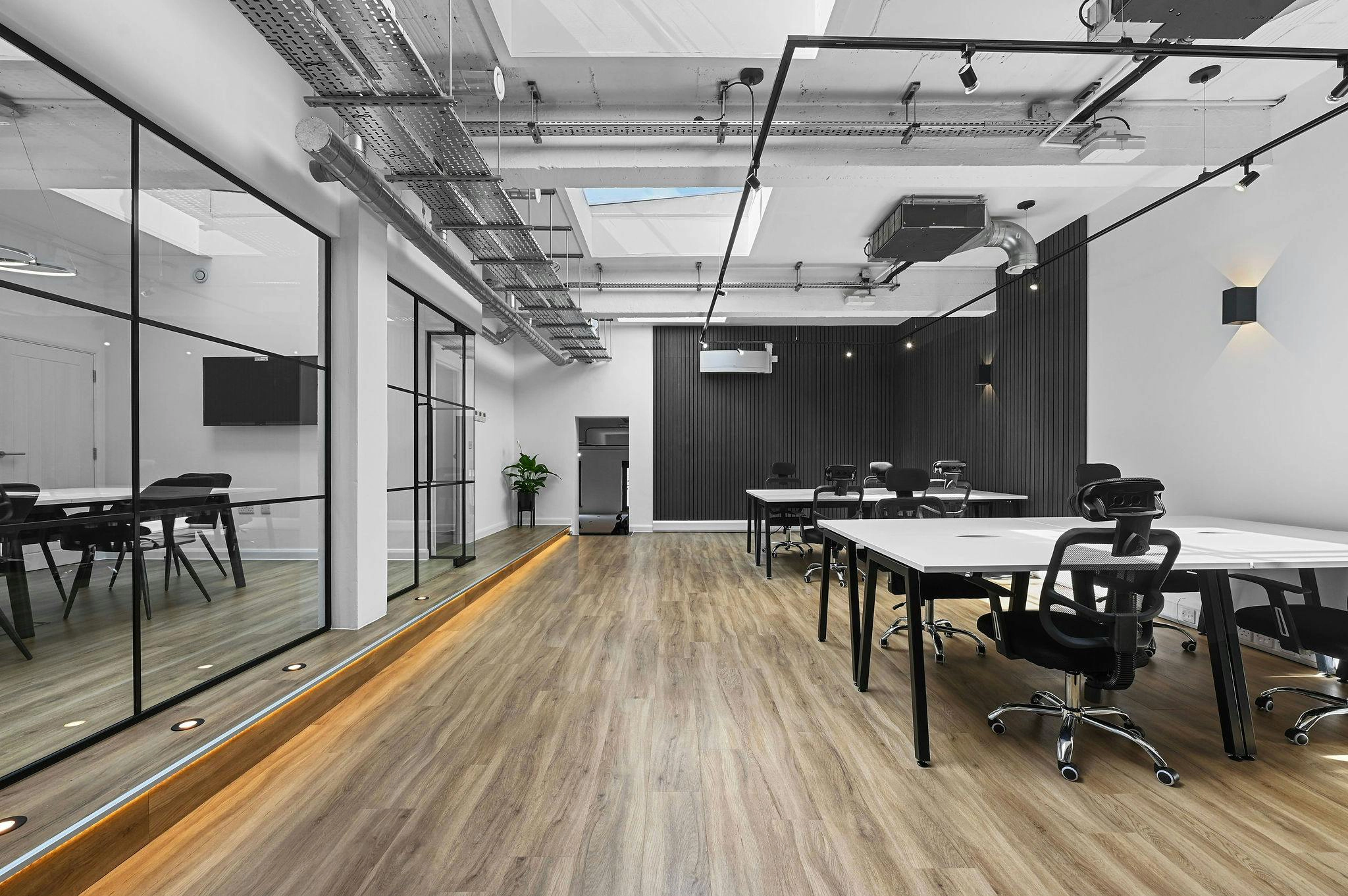 Modern office space with wooden flooring, white desks, black ergonomic chairs, and a glass-walled meeting room on the left.