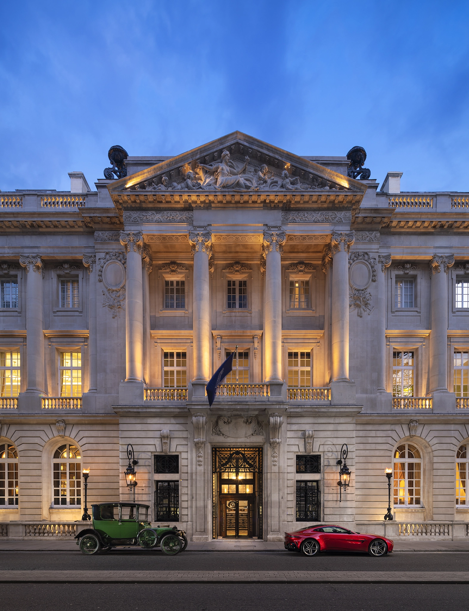 Illuminated classic stone building facade with columns and sculptures at dusk, flanked by a vintage green car on the left and a modern red sports car on the right.