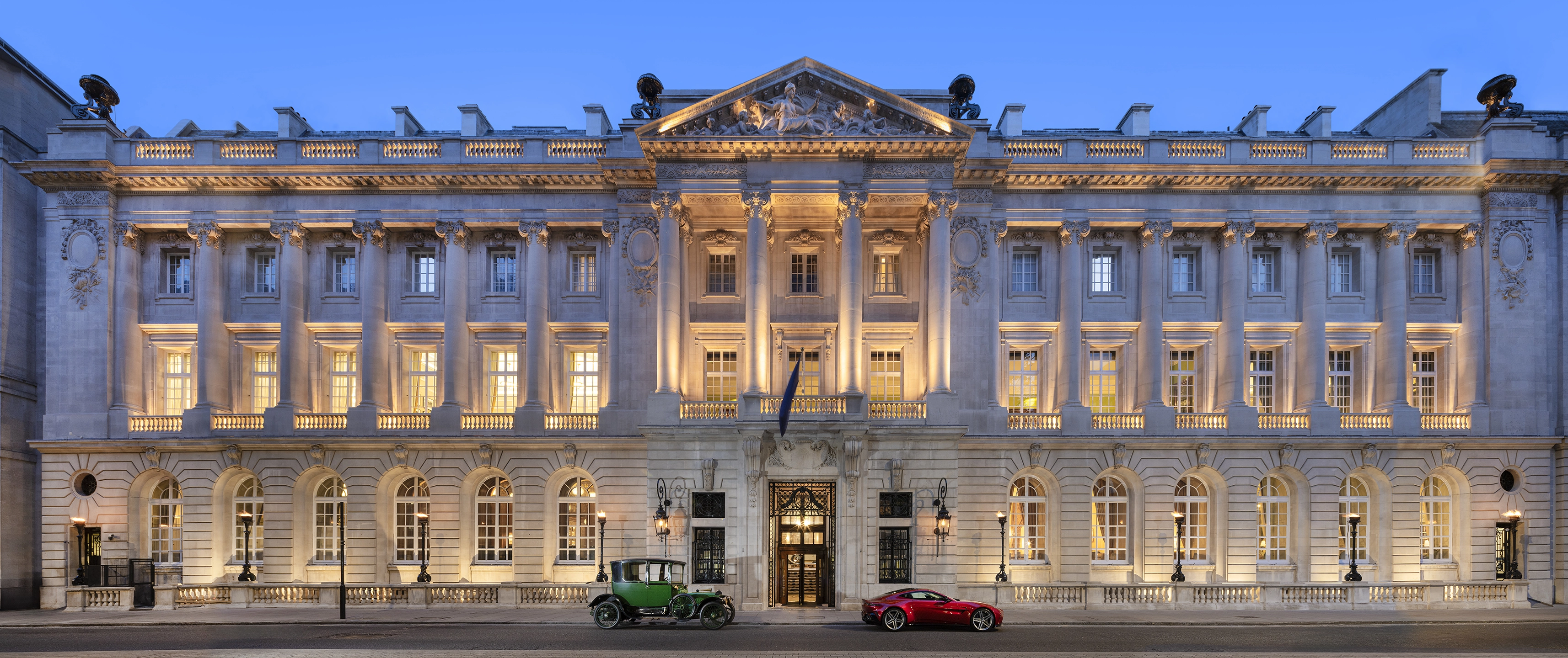 Grand neoclassical building facade lit warmly at dusk with vintage green car and modern red sports car parked in front.