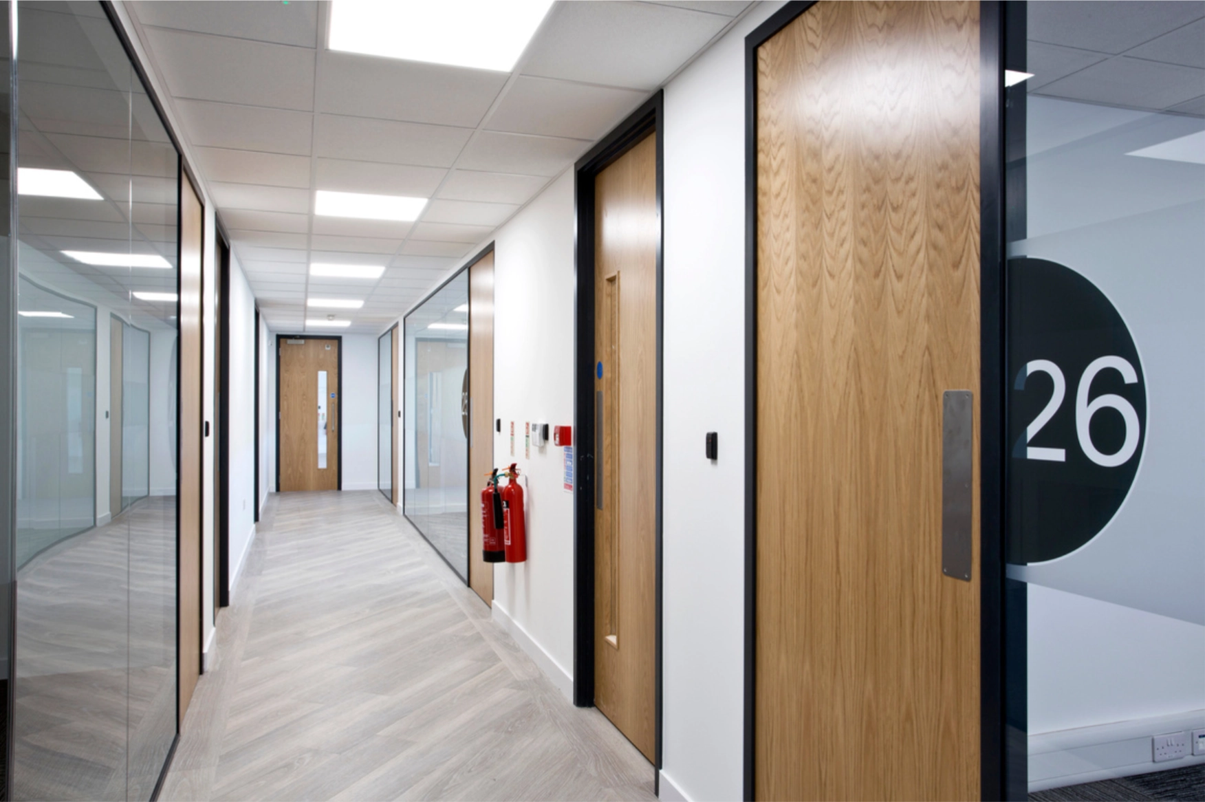 Modern office corridor with glass walls, wooden doors, and two red fire extinguishers mounted on white wall.