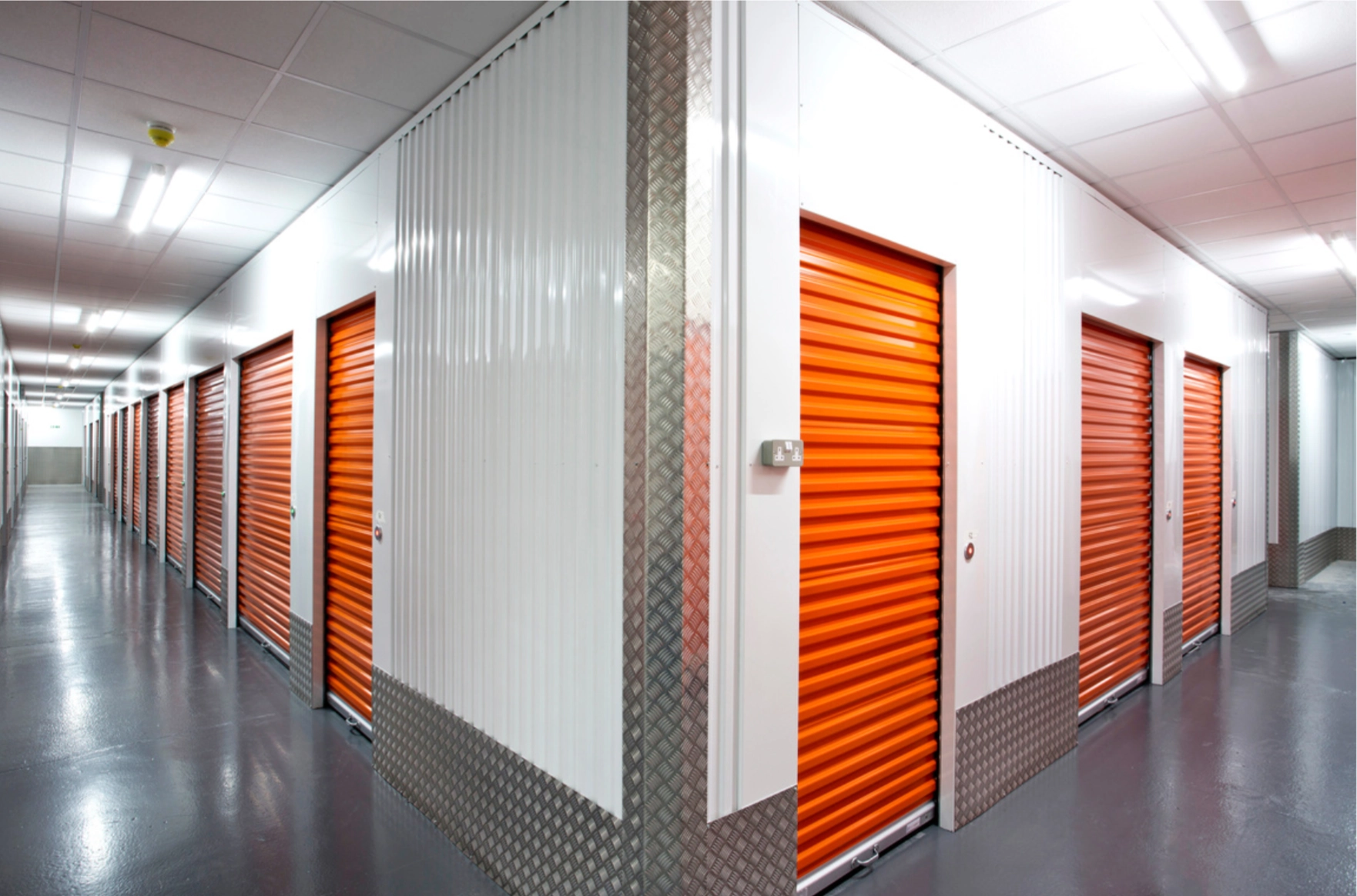 Interior hallway of a self-storage facility with orange roll-up doors on either side and a polished concrete floor.